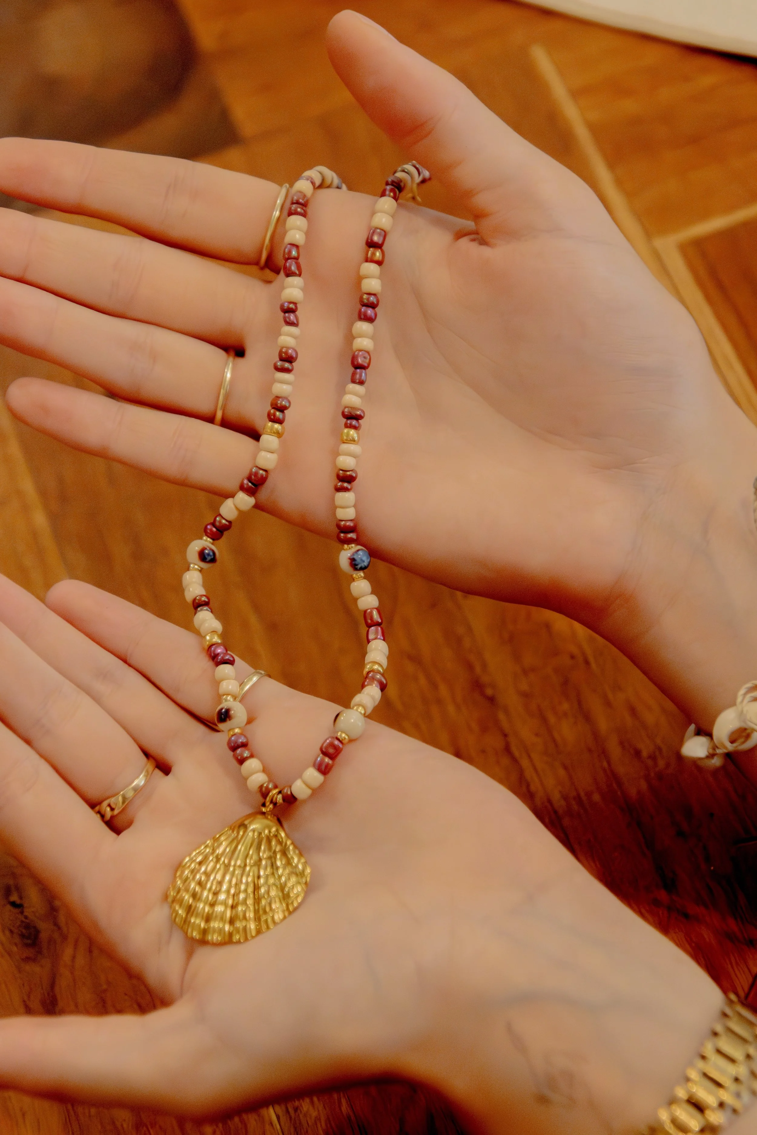 A person's hands displaying a beaded necklace with a gold seashell pendant. The necklace has cream and red beads with some dark blue beads featuring images, and the person is wearing multiple rings and bracelets.