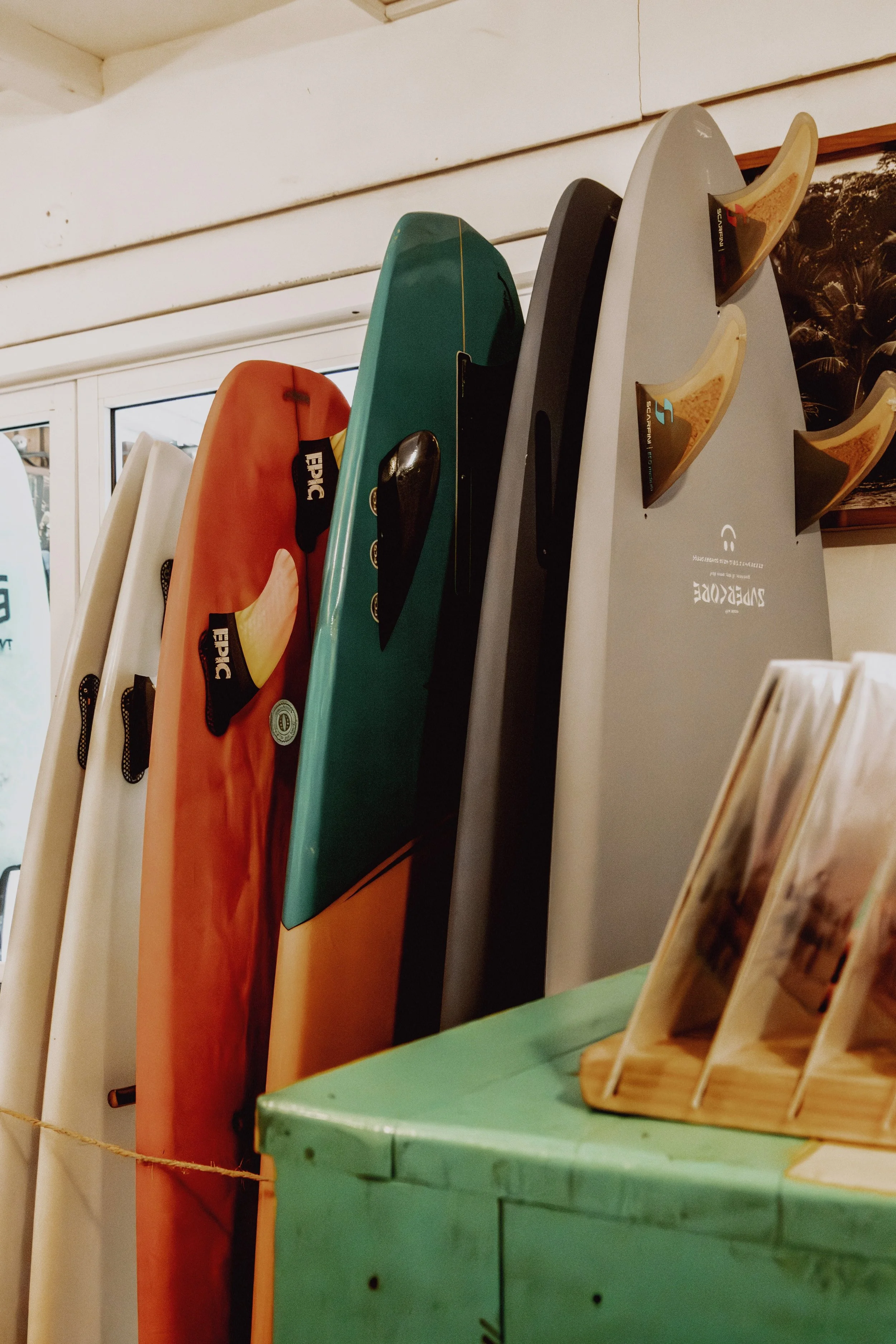 Multiple surfboards in various colors, including white, orange, teal, black, and gray, standing upright indoors next to a window and a green counter with a wooden brochure holder on top.