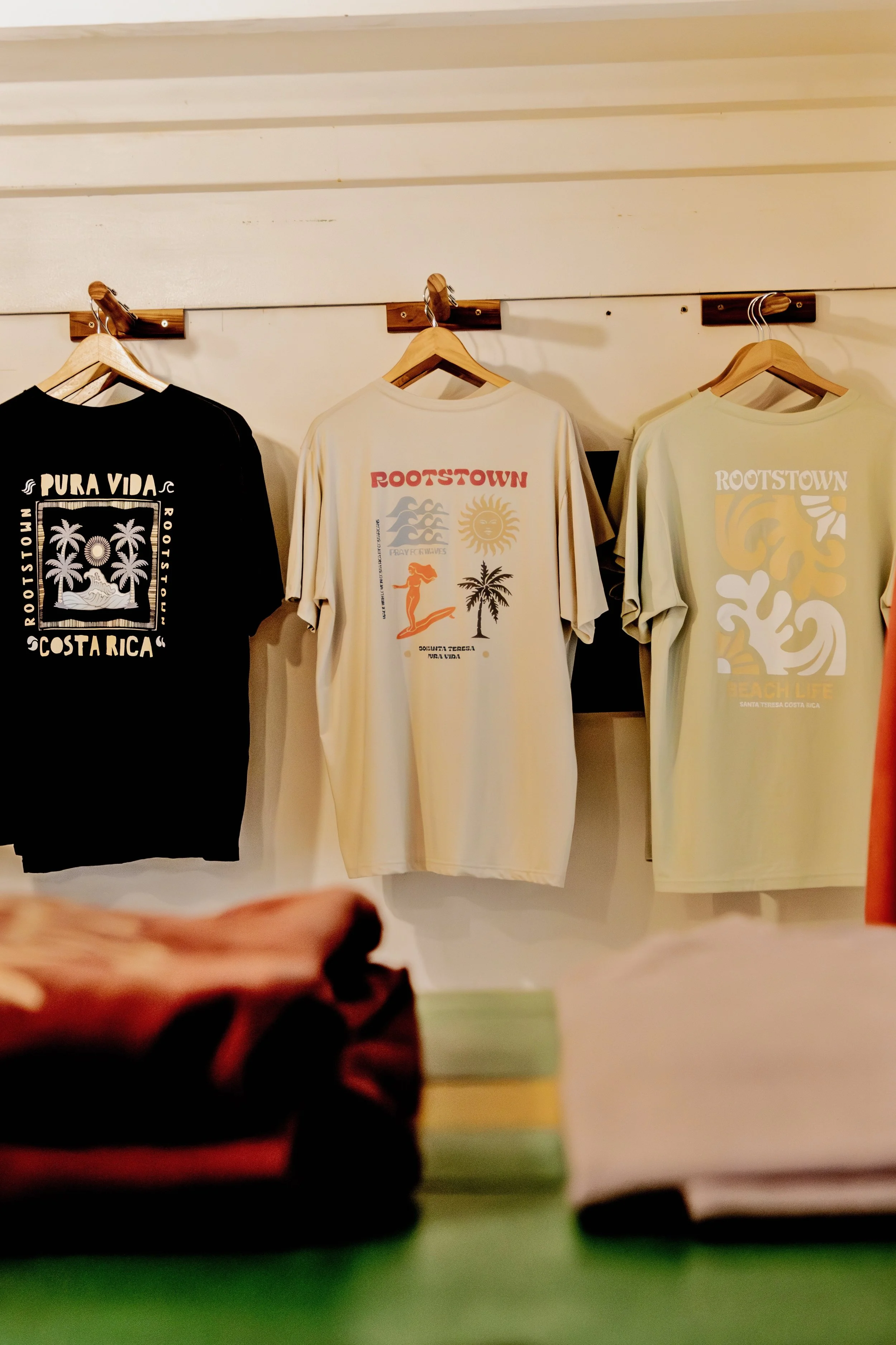Three T-shirts hanging on a wall display, featuring tropical and beach-themed designs related to Costa Rica and Santa Teresa, with folded clothes on a table in the foreground.