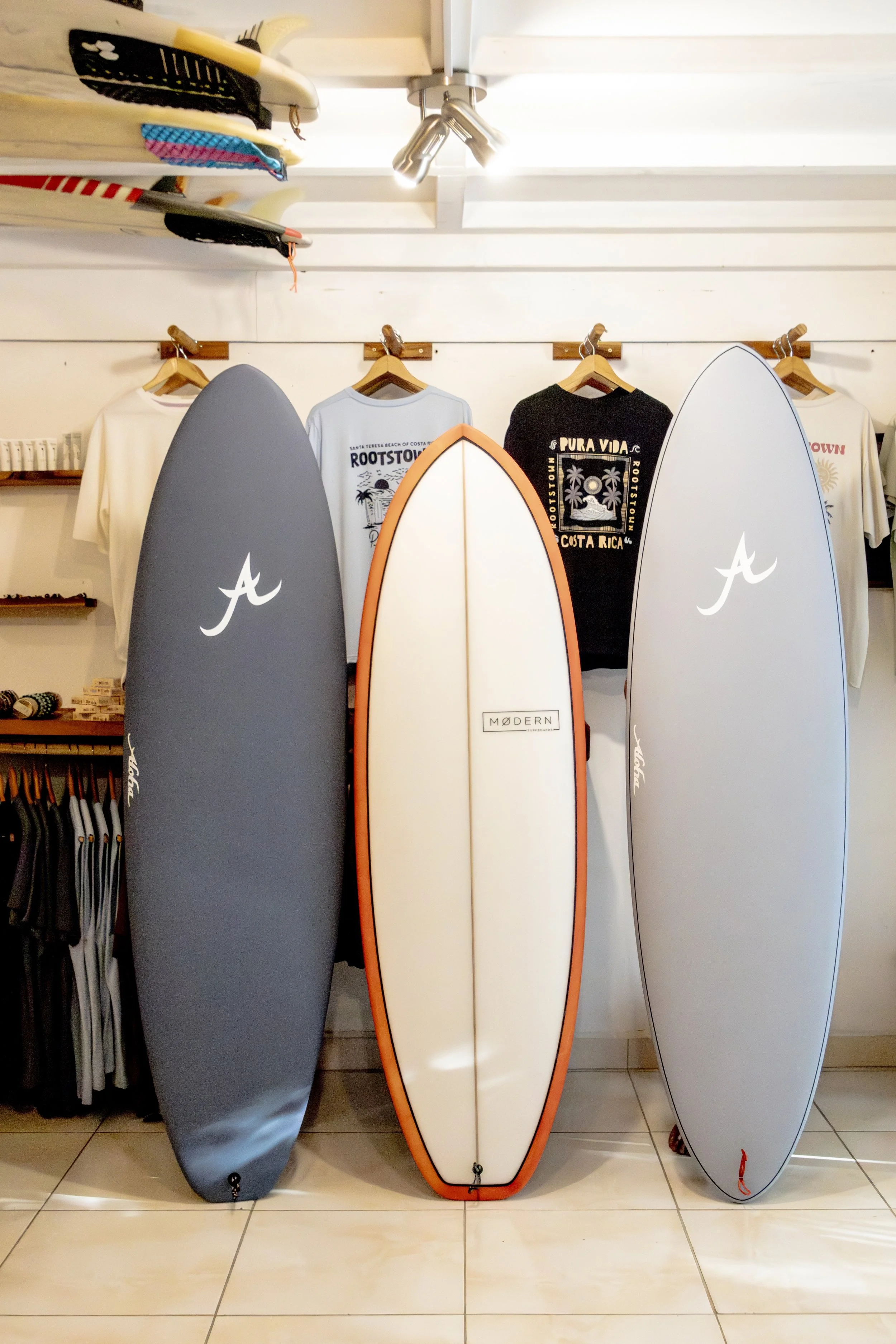 Three surfboards standing upright on a tiled floor inside a store. The surfboards are gray, cream with a red border, and light gray. Behind them are t-shirts hanging on a wall, with surf-themed graphics and text.