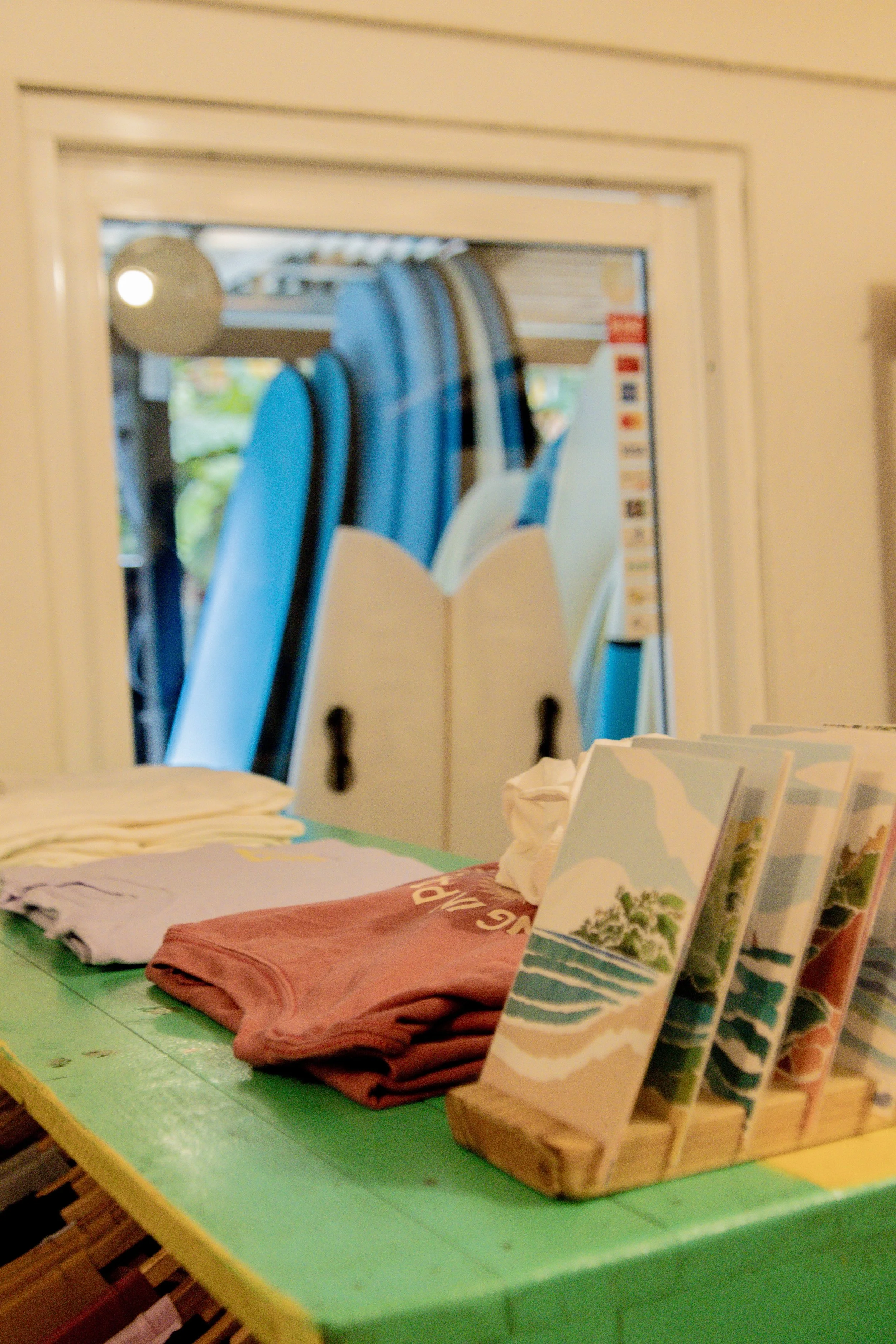 A display table with folded clothes and small beach-themed artwork in the foreground, with surfboards in a rack behind a mirror in the background.