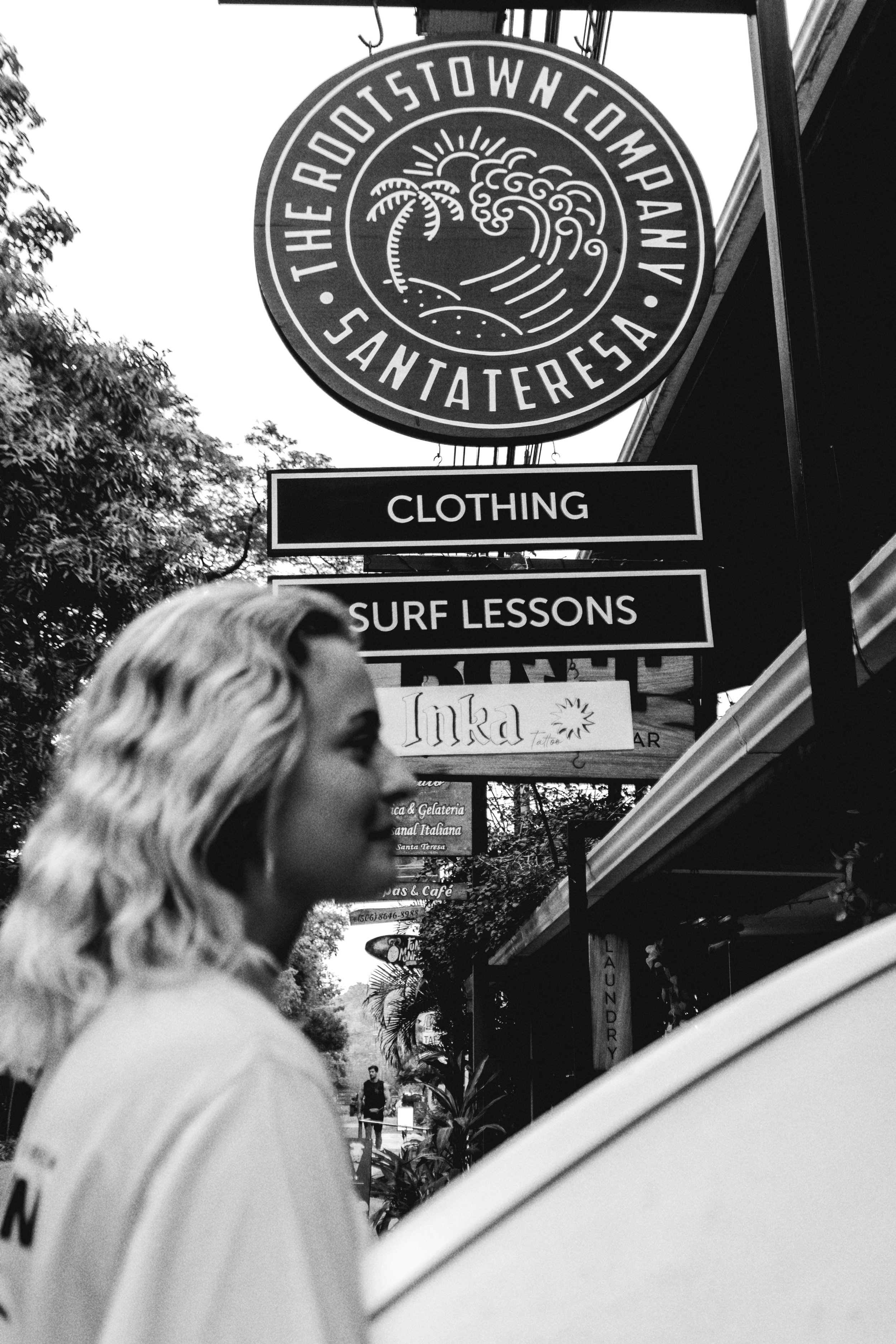 Black and white photo of a woman with wavy blonde hair standing on a street sidewalk, with various shop signs above including "Clothing," "Surf Lessons," and "Inka" in the background.