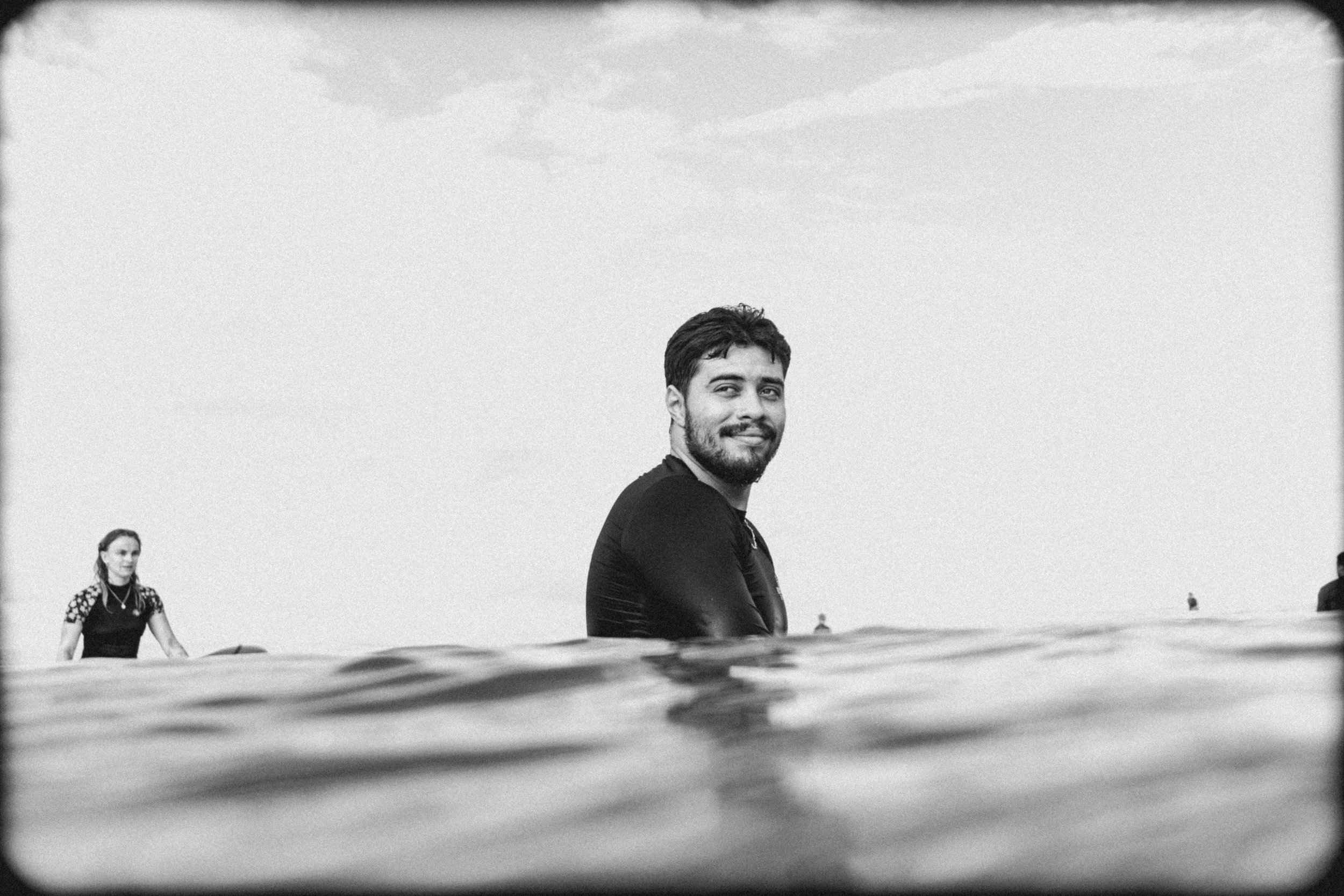 Black and white photo of a man smiling at the camera, partially submerged in water, with a woman in the background also in the water, and a cloudy sky above.