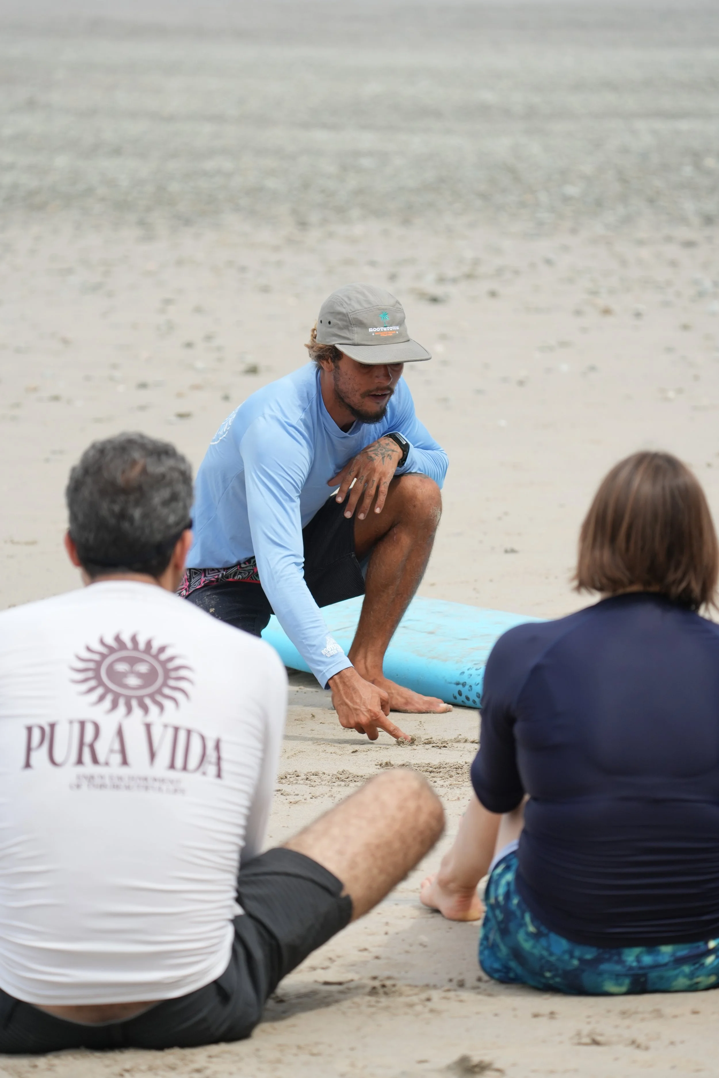 Group of people sitting on the sand at the beach, listening to a man in a light blue shirt and gray hat, who is kneeling and explaining something with a surfboard nearby.