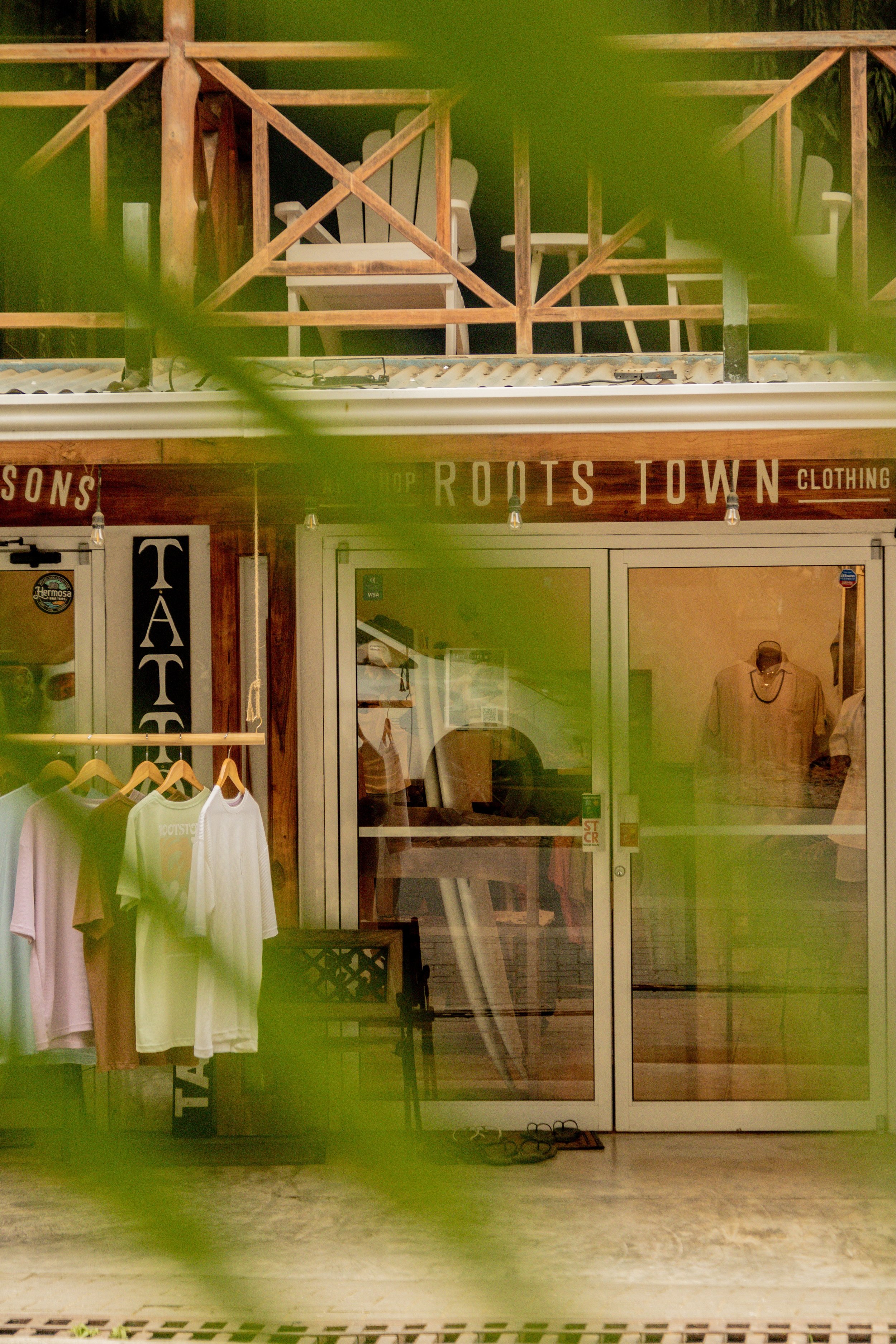 Storefront with clothing display, wooden balcony with chairs on upper level, and greenery partially obscuring view.
