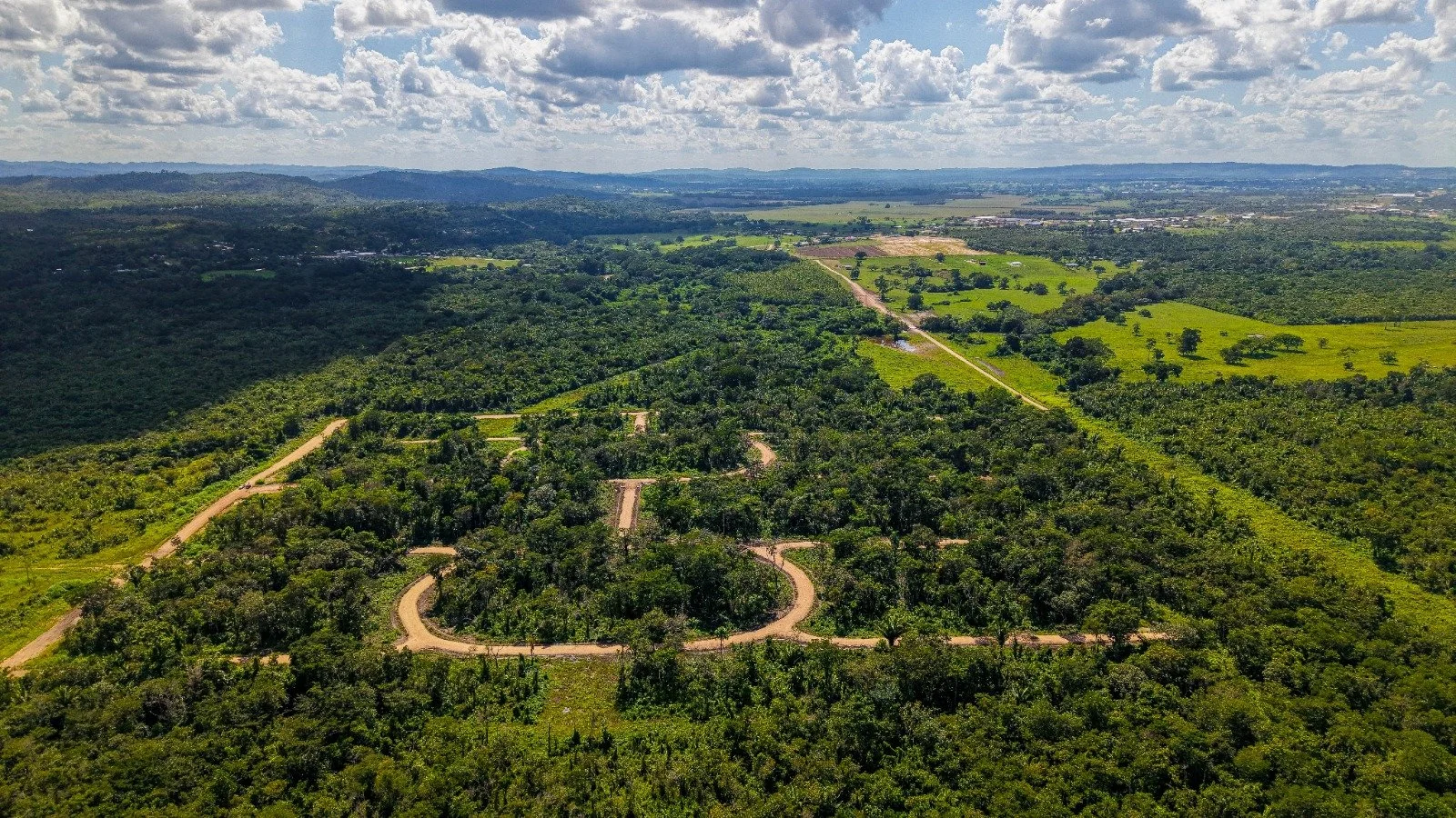 Aerial view of a landscape with lush green forests, winding dirt roads, scattered trees, and distant hills under a partly cloudy sky.