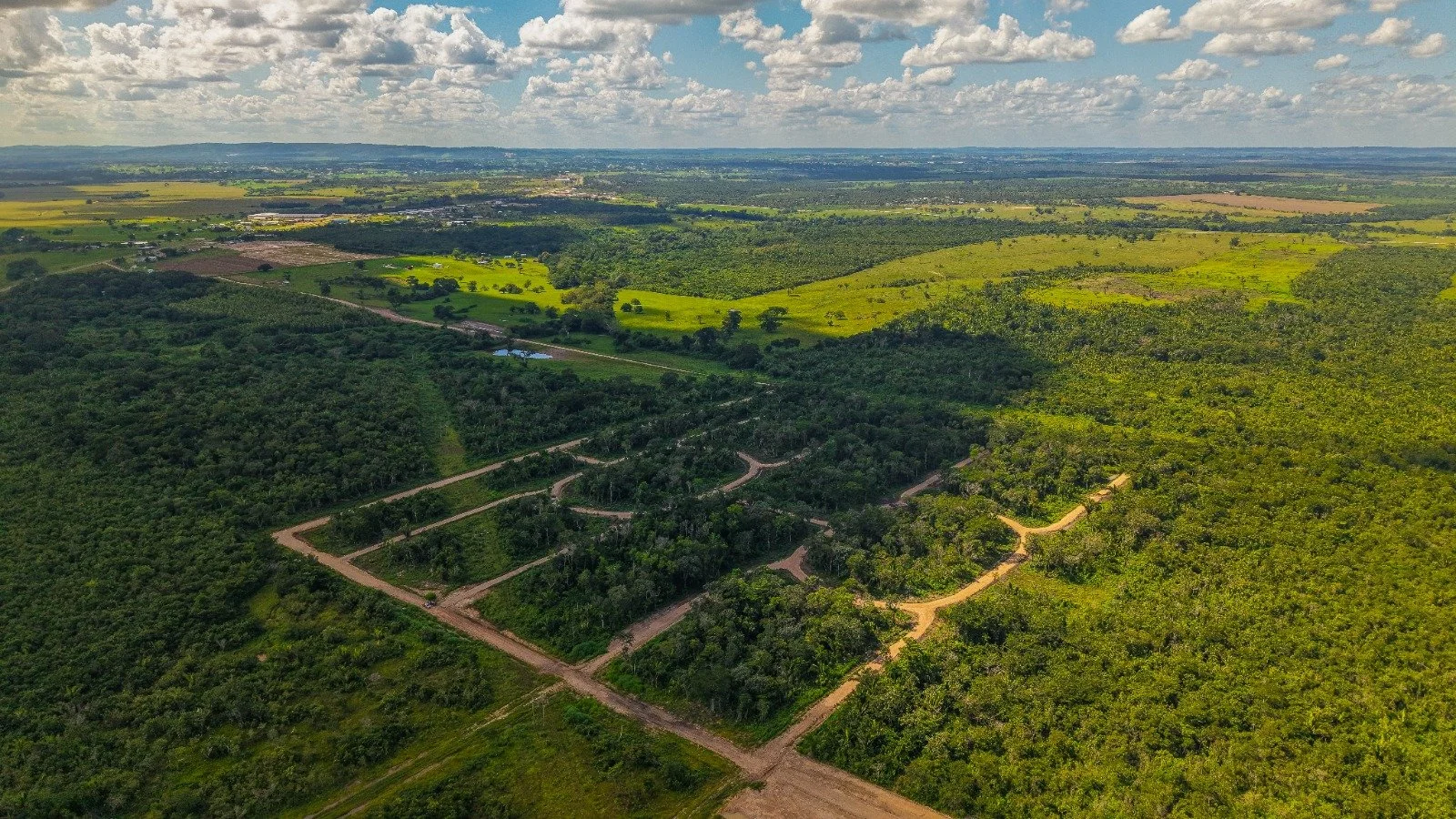 Aerial view of a vast rural landscape with green fields, patches of forest, and dirt roads under a partly cloudy sky.