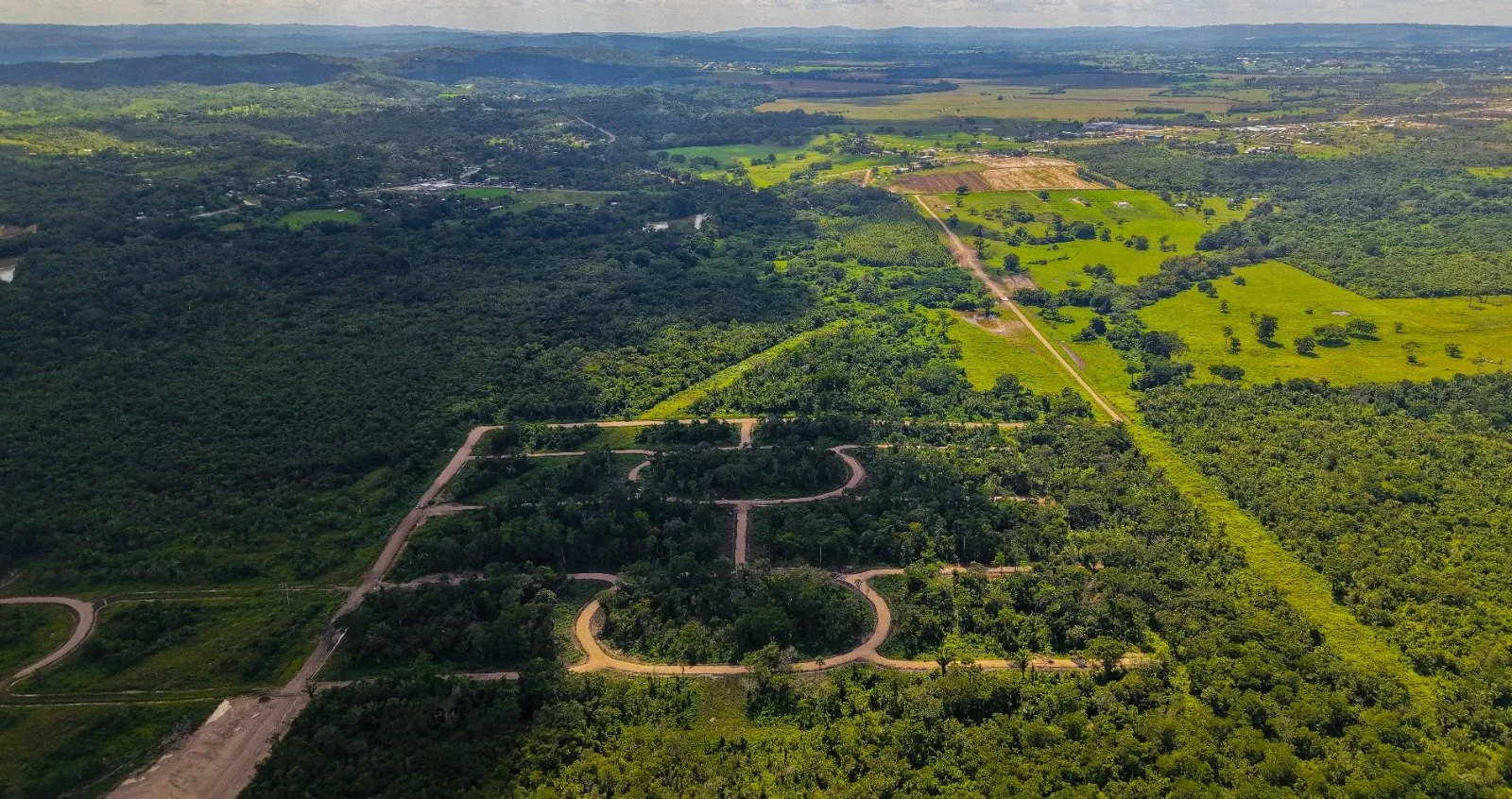 Aerial view of a green, rural landscape with winding roads, dense forests, grassy fields, and some distant distant buildings and hills.
