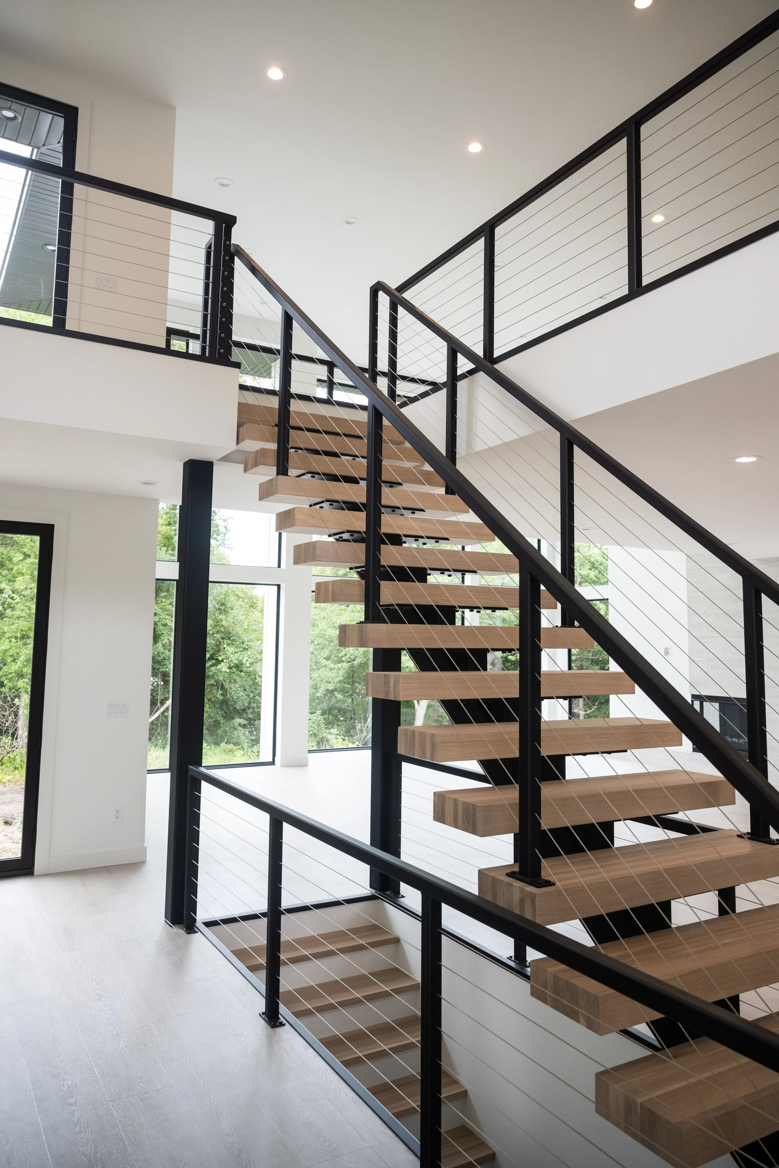 Modern staircase with wooden steps and black metal railings inside a house with large windows and white walls.
