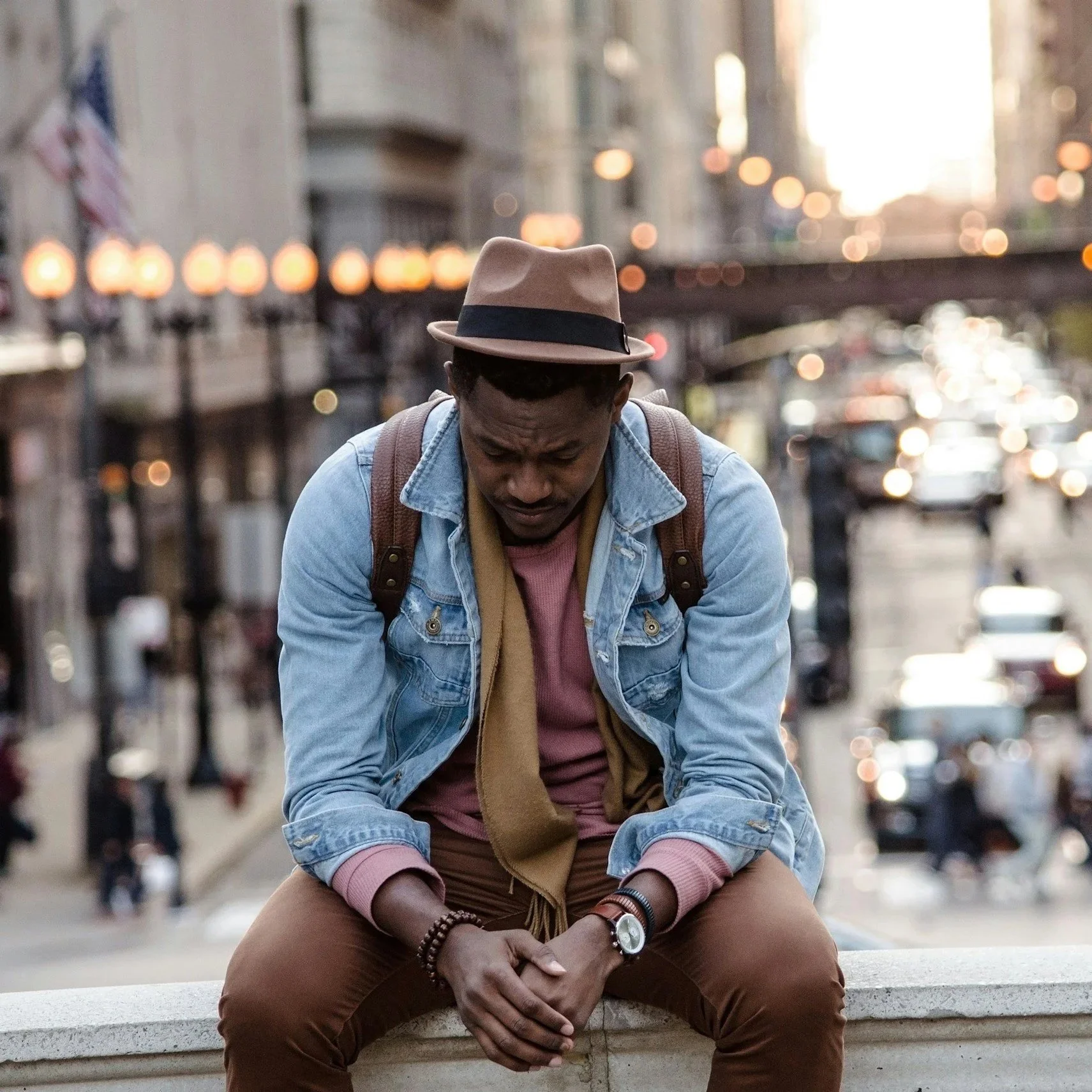 A man sitting on a ledge with a city street in the background, looking down with a thoughtful expression.
