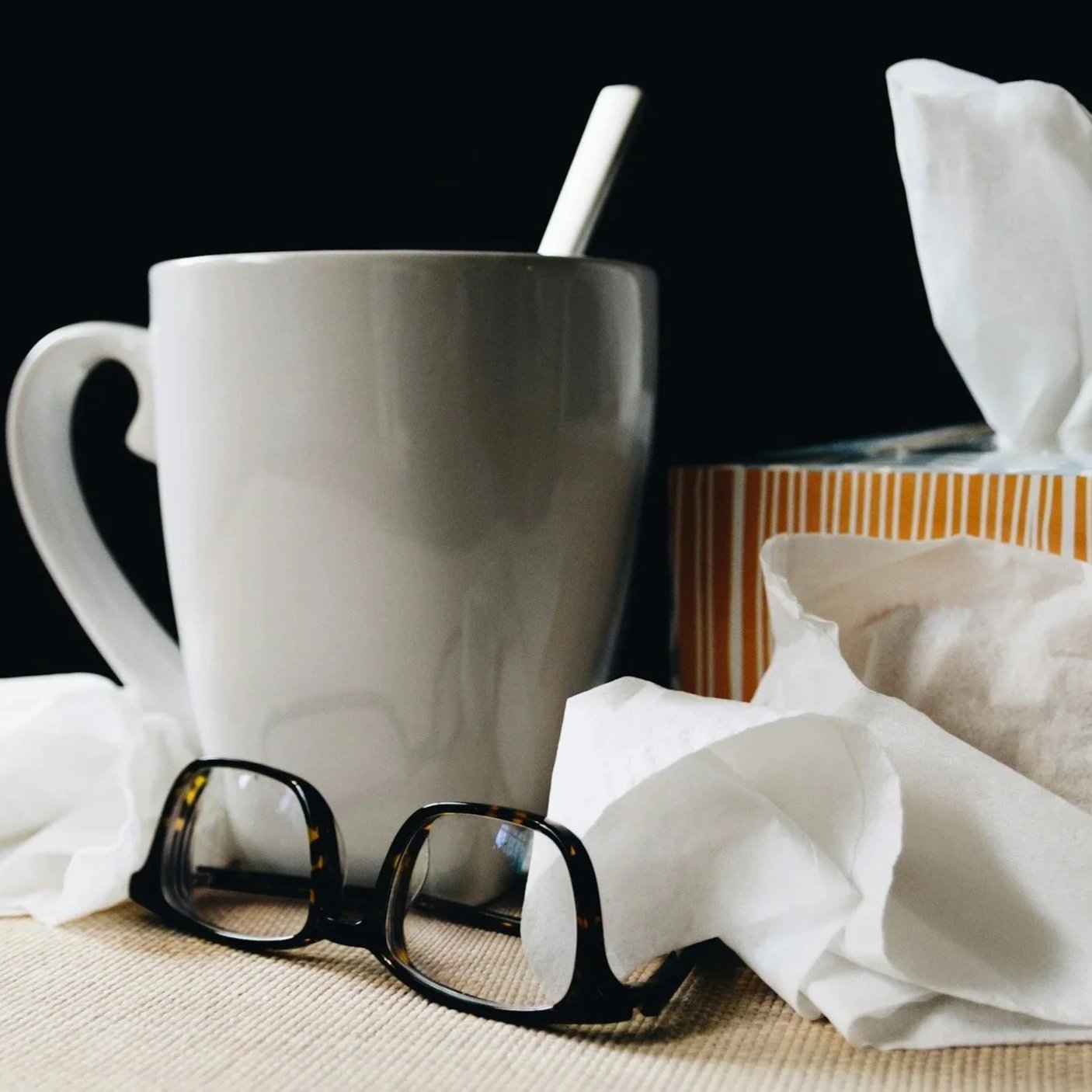 A white coffee mug with a spoon, glasses, crumpled tissue papers, and a tissue box on a table against a black background.