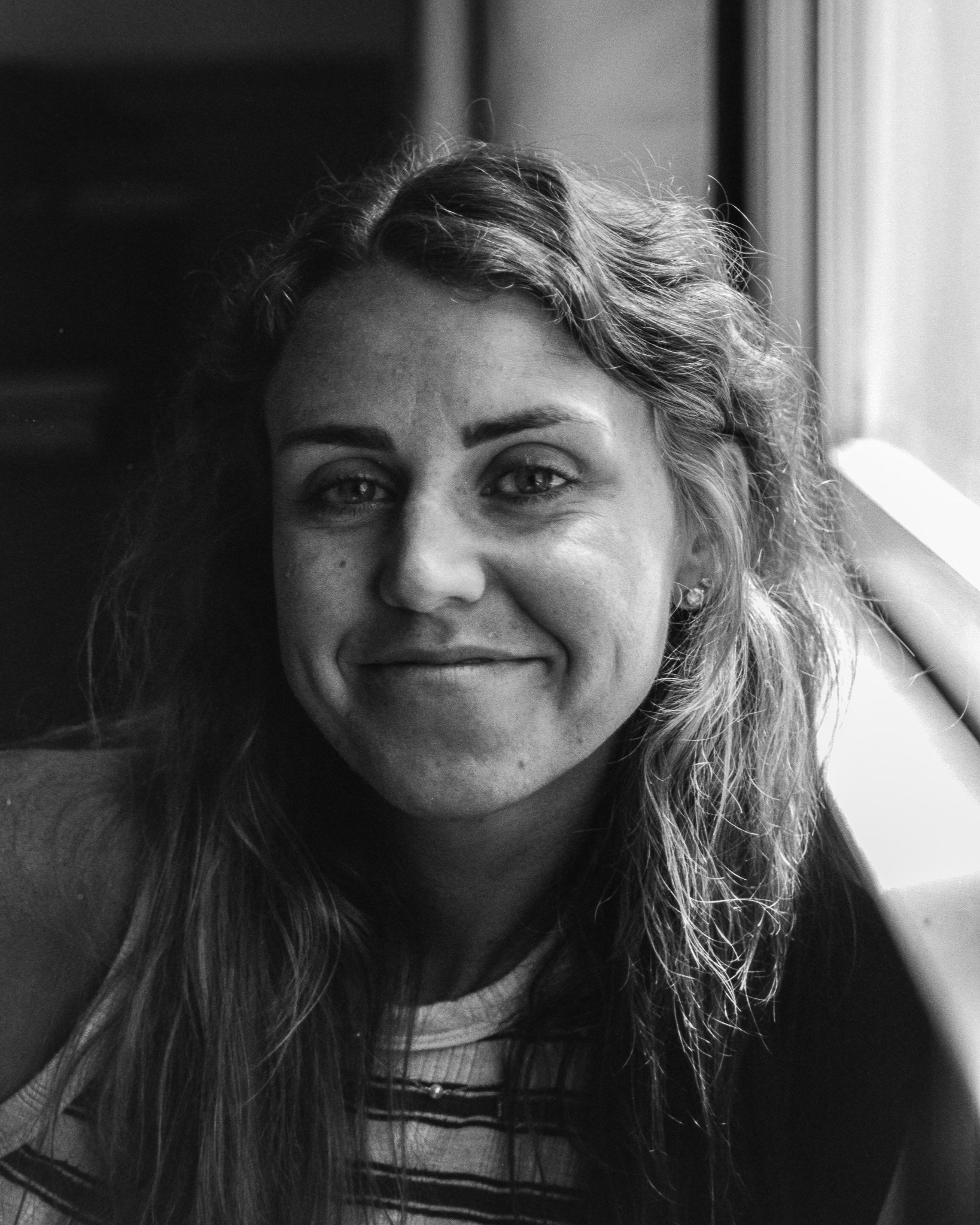 A black-and-white photograph of a woman with wavy hair smiling, looking at the camera, seated by a window.