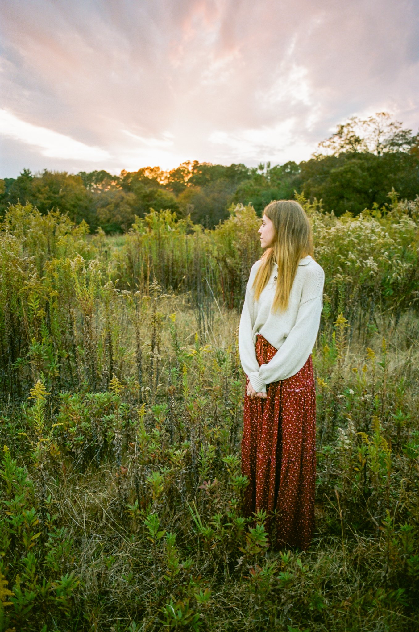 A young woman with long blonde hair standing in a field of tall grass and wild plants during sunset, with trees in the background and a partly cloudy sky.