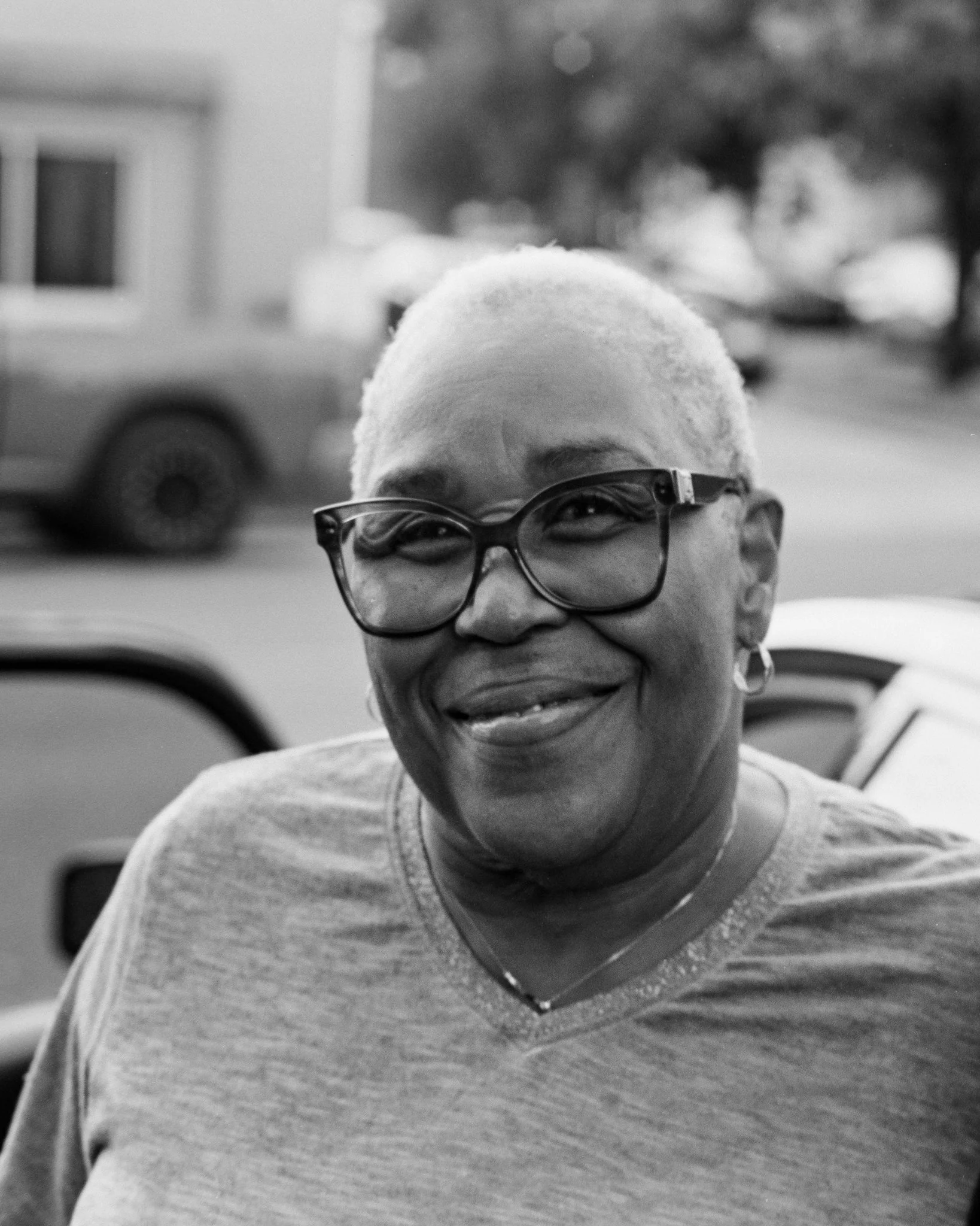 A smiling woman with short, light-colored hair, glasses, and earrings, standing outdoors near a parked car.