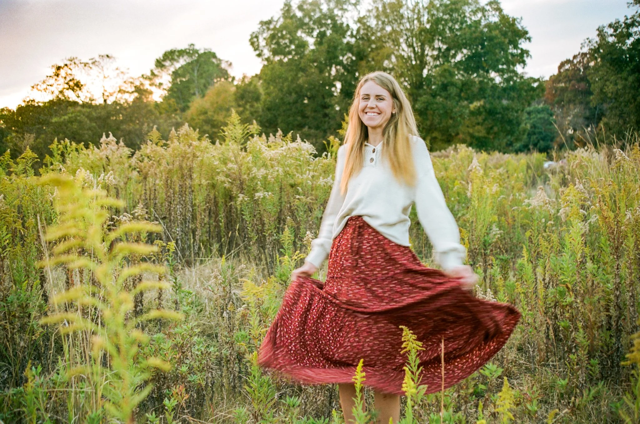 Young woman smiling in a field of tall grass and wildflowers at sunset, wearing a white sweater and a long red skirt.