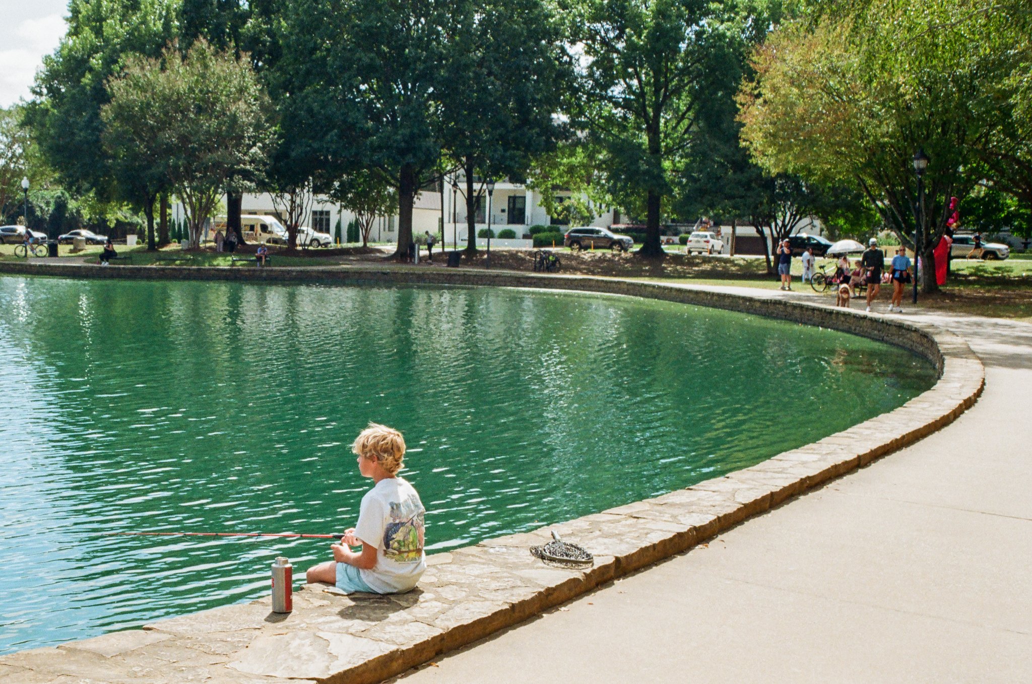 A young boy with blonde hair sitting on a stone ledge by a pond, fishing with a rod. The pond is surrounded by park greenery and trees, with people walking and biking in the background.