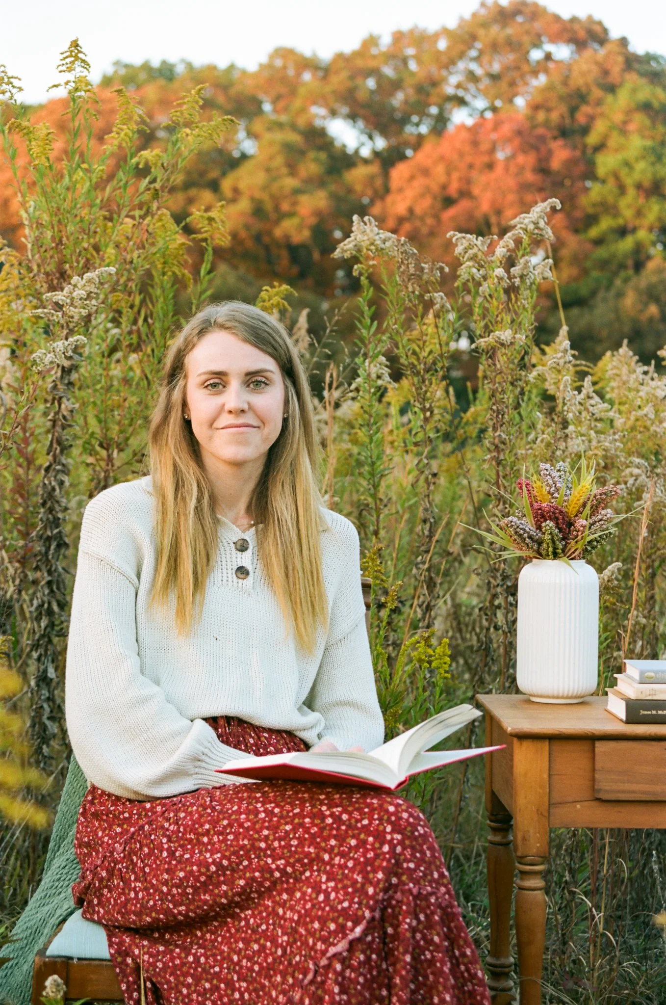 A young woman with long, light brown hair sitting outdoors surrounded by autumn foliage. She is wearing a white sweater with dark buttons and a red floral skirt, holding an open book on her lap. Next to her is a wooden table with a white vase holding