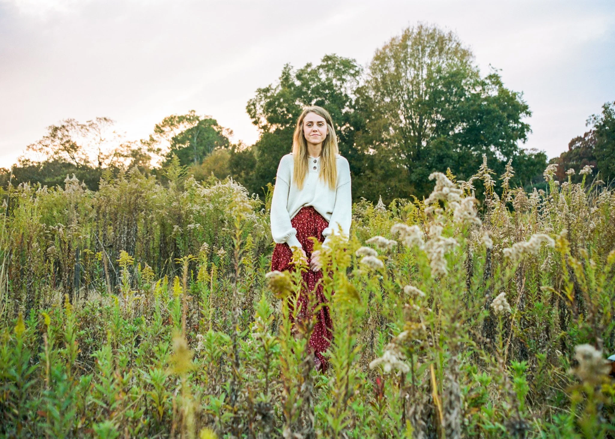 A woman with long blonde hair standing in a field of tall plants with trees and a cloudy sky in the background.