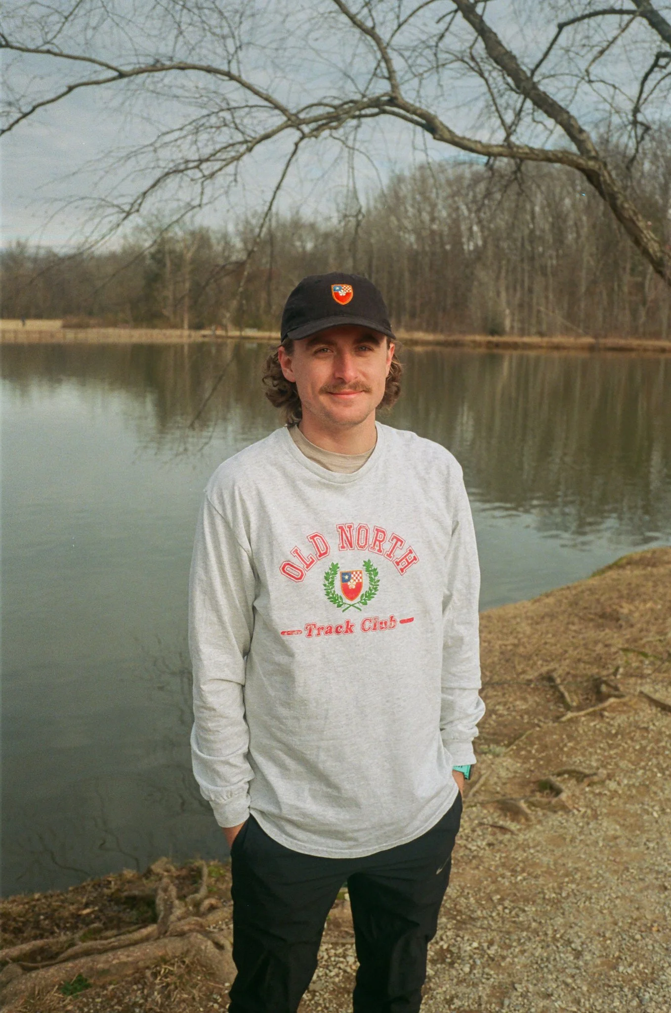 A man standing outdoors near a body of water, wearing a black cap and a gray hoodie with 'Old North Track Club' written on it. He has brown hair, a mustache, and is smiling.