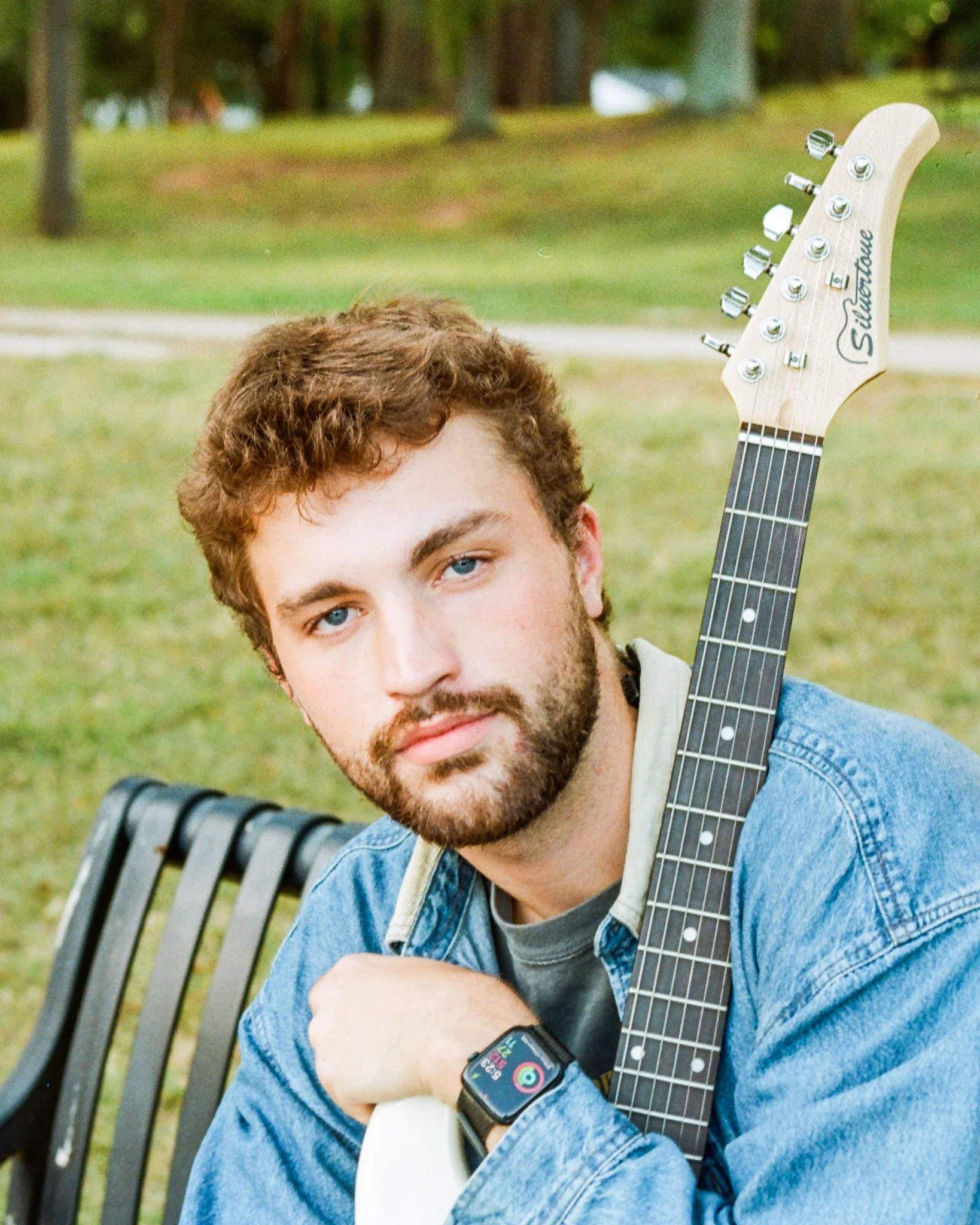 A young man with brown curly hair and blue eyes sitting on a park bench, holding a white electric guitar with the headstock visible and the brand name Silhouette, wearing a denim jacket and a smartwatch, with trees and grass in the background.