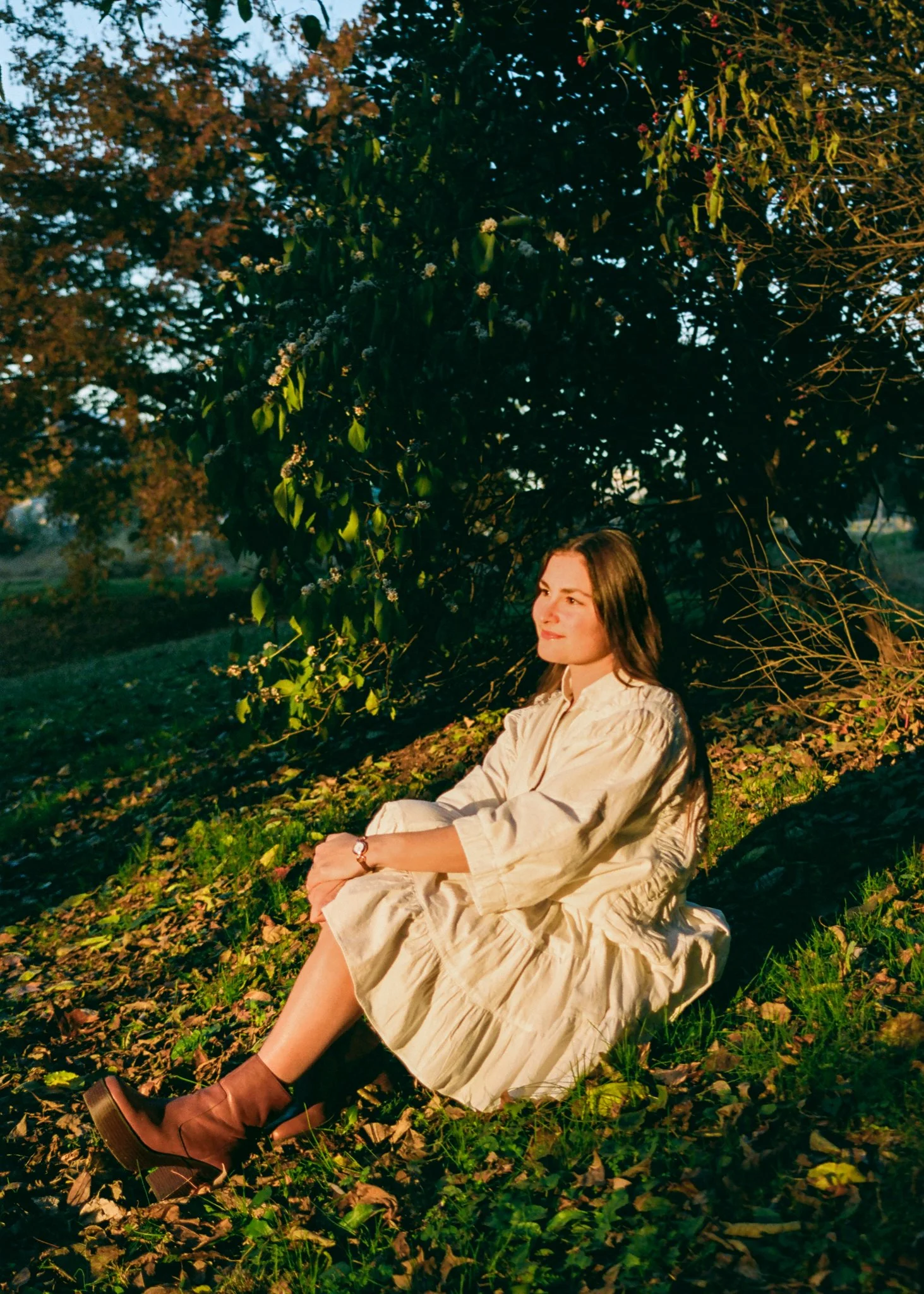 A woman sitting on the grass in a park during sunset, surrounded by green foliage and fallen leaves, wearing a beige dress and brown boots, with sunlight illuminating her face.