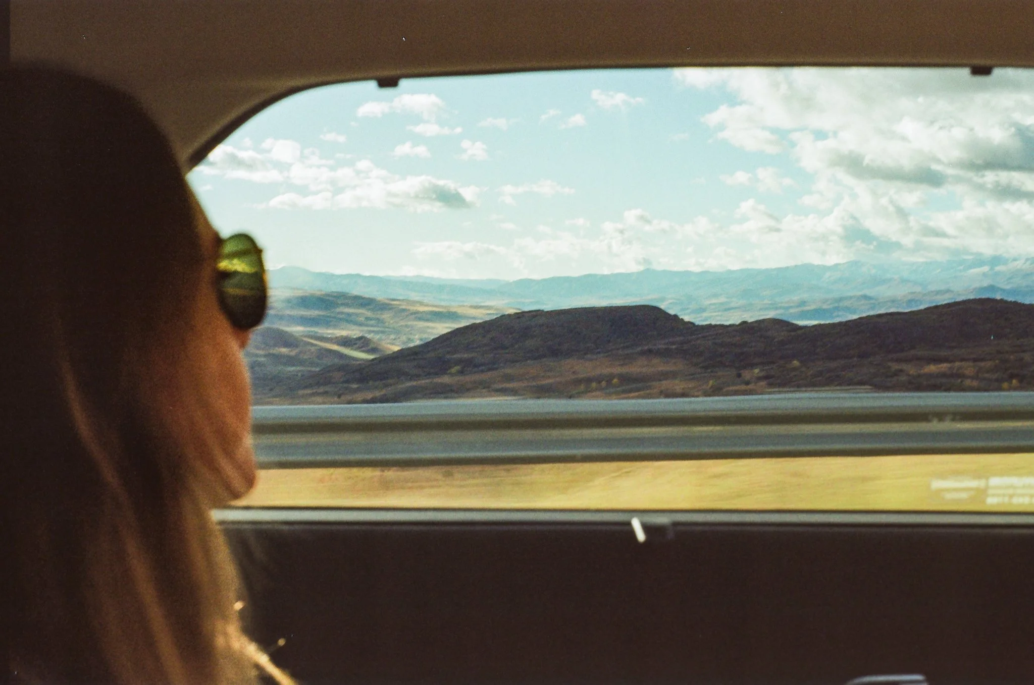 A woman with red hair and sunglasses sitting in a car, looking out the window at a landscape of mountains and hills under a partly cloudy sky.