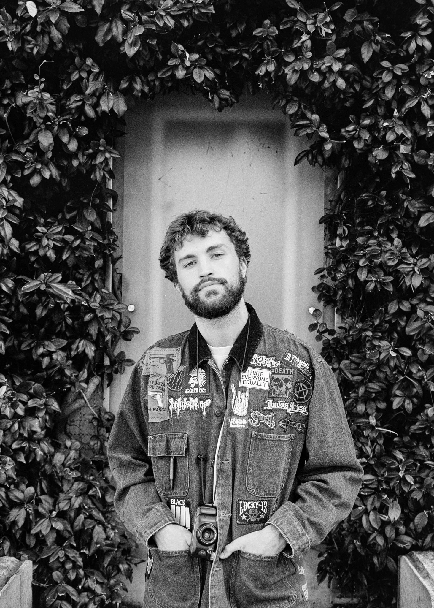 A young man with curly hair and a beard standing in front of a door, framed by dense foliage, wearing a patched denim jacket and holding a camera around his neck.