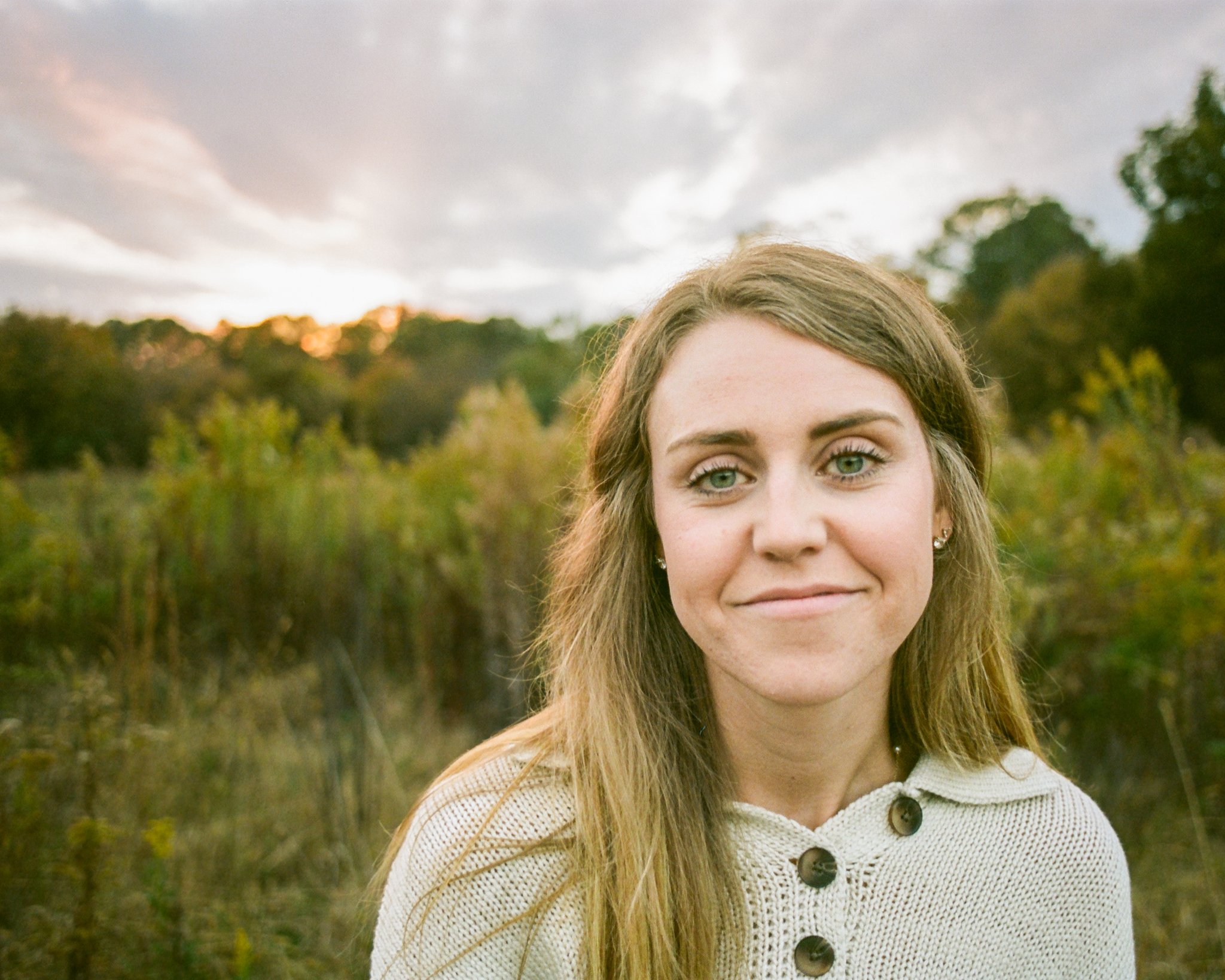 A young woman with long, light brown hair and light blue eyes smiling outdoors during sunset, wearing a cream-colored sweater with large buttons.