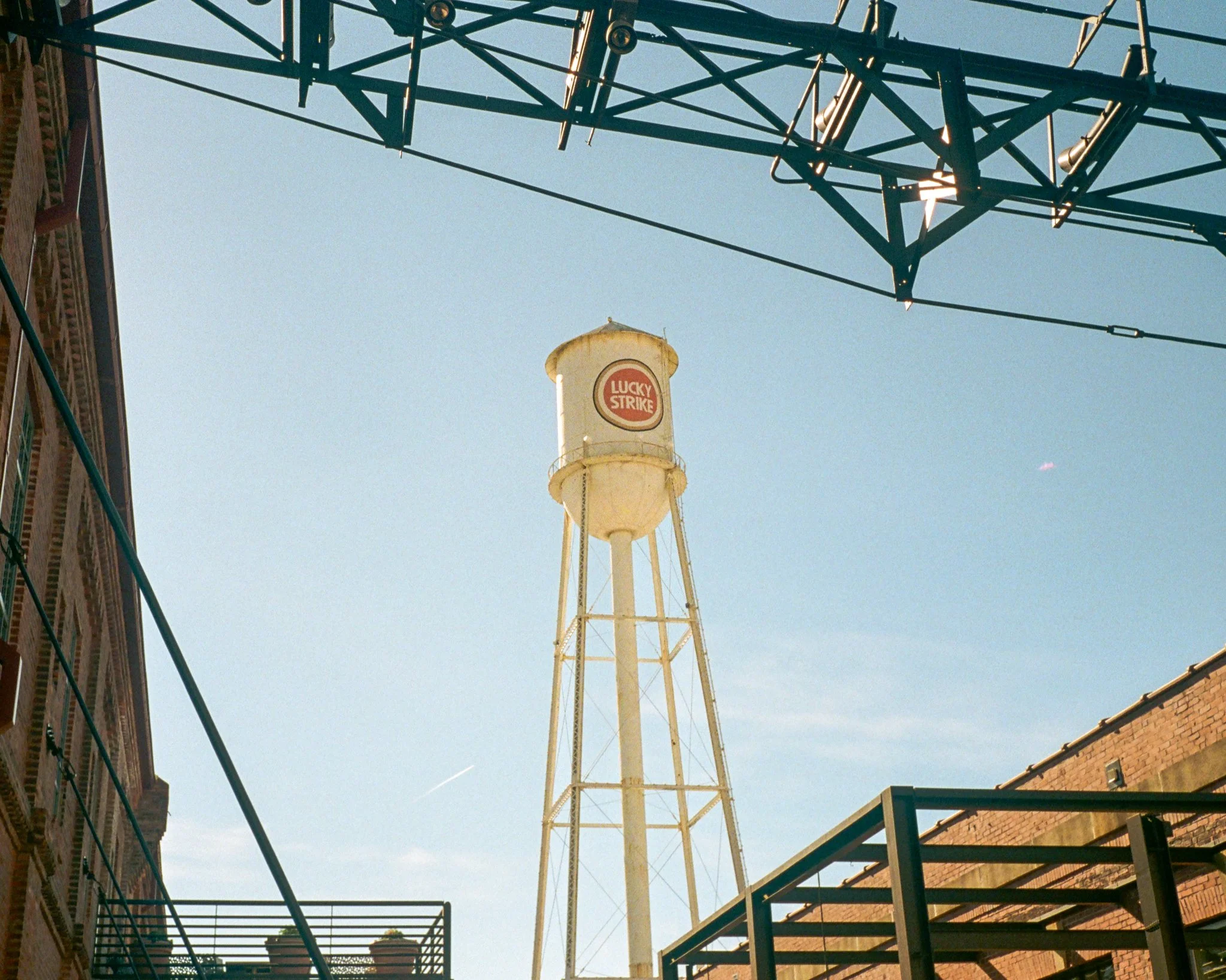 An old water tower with a Lucky Strike logo painted on it, situated between brick and metal buildings, with a clear blue sky above.