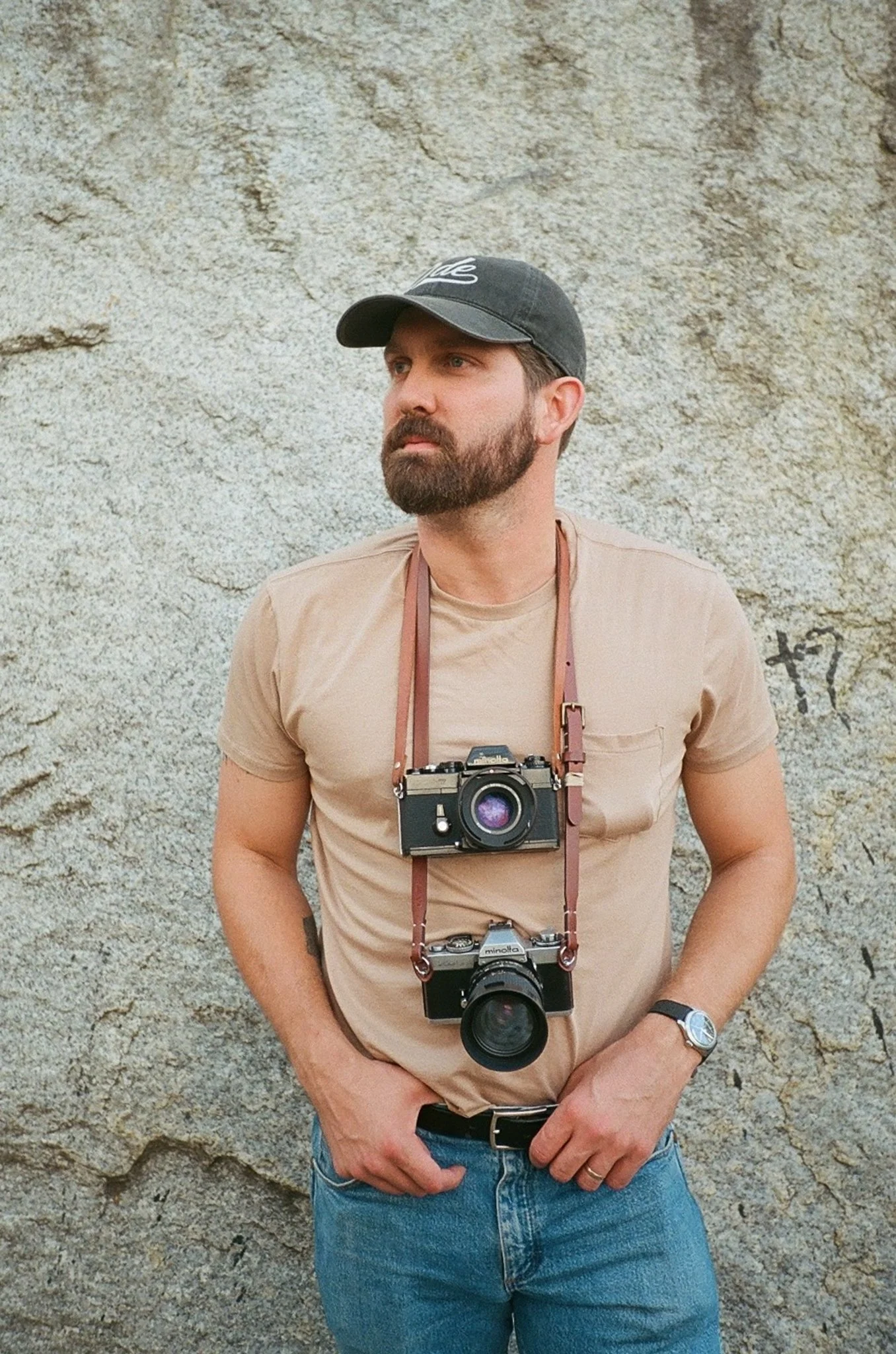 A man with a beard wearing a black cap, a beige t-shirt, and jeans, standing against a textured concrete wall with graffiti, carrying two cameras around his neck and accessories.
