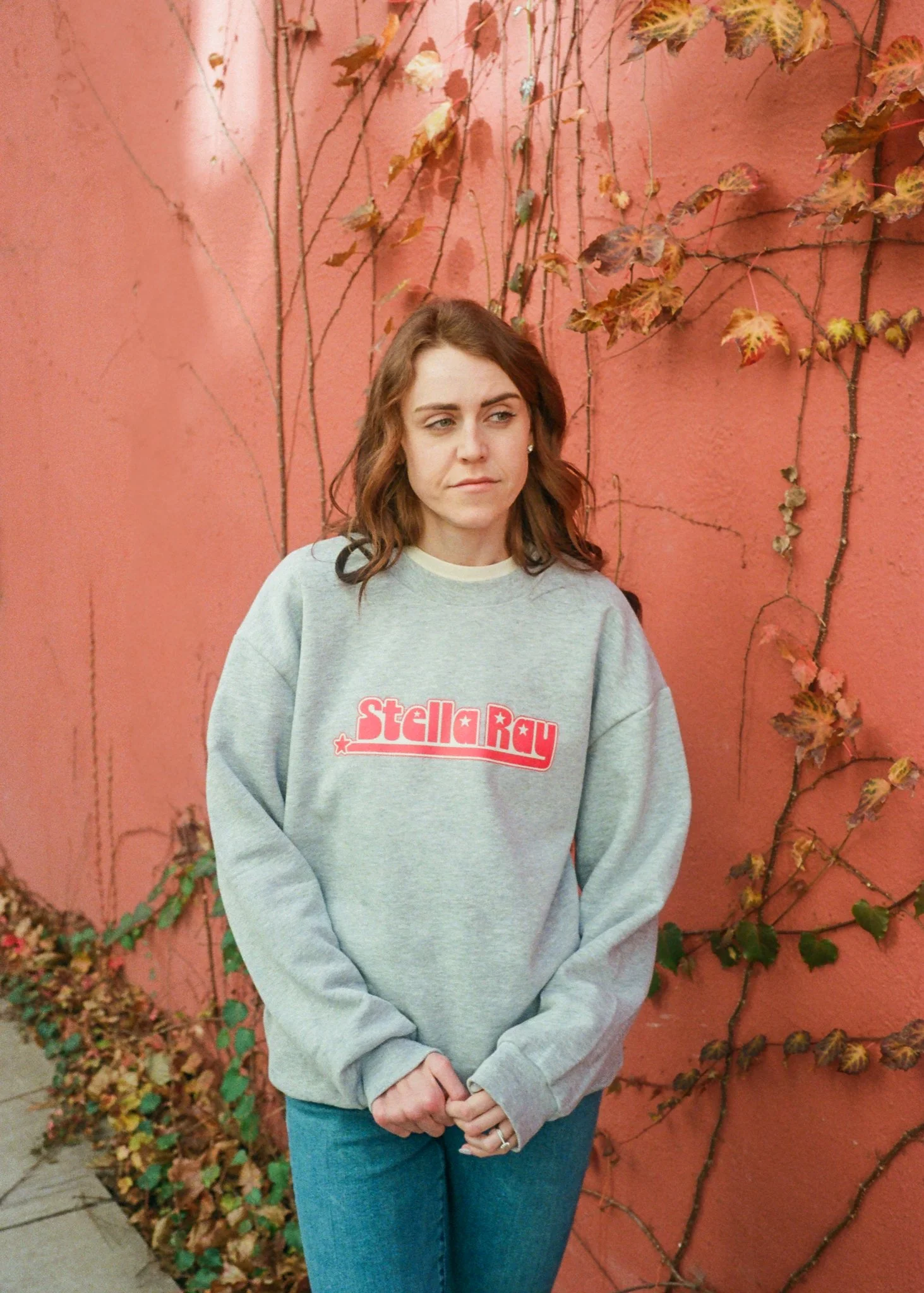 A woman in a gray sweatshirt with 'StellaRay' written on it, standing against a pink wall covered with dry and green vines.