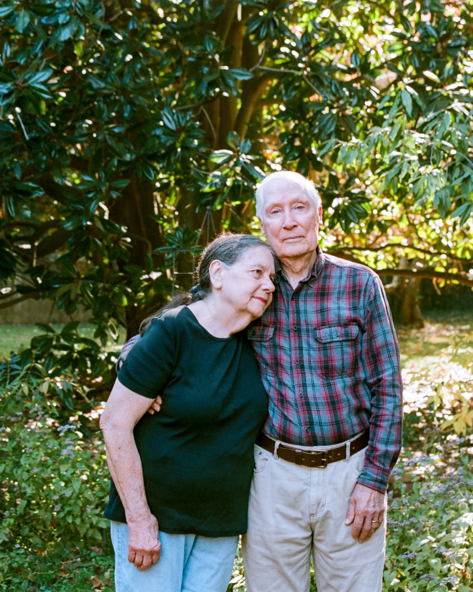 An elderly man and woman standing close together outdoors, with the woman leaning her head on the man's shoulder. They are surrounded by green foliage and sunlight filtering through the trees.