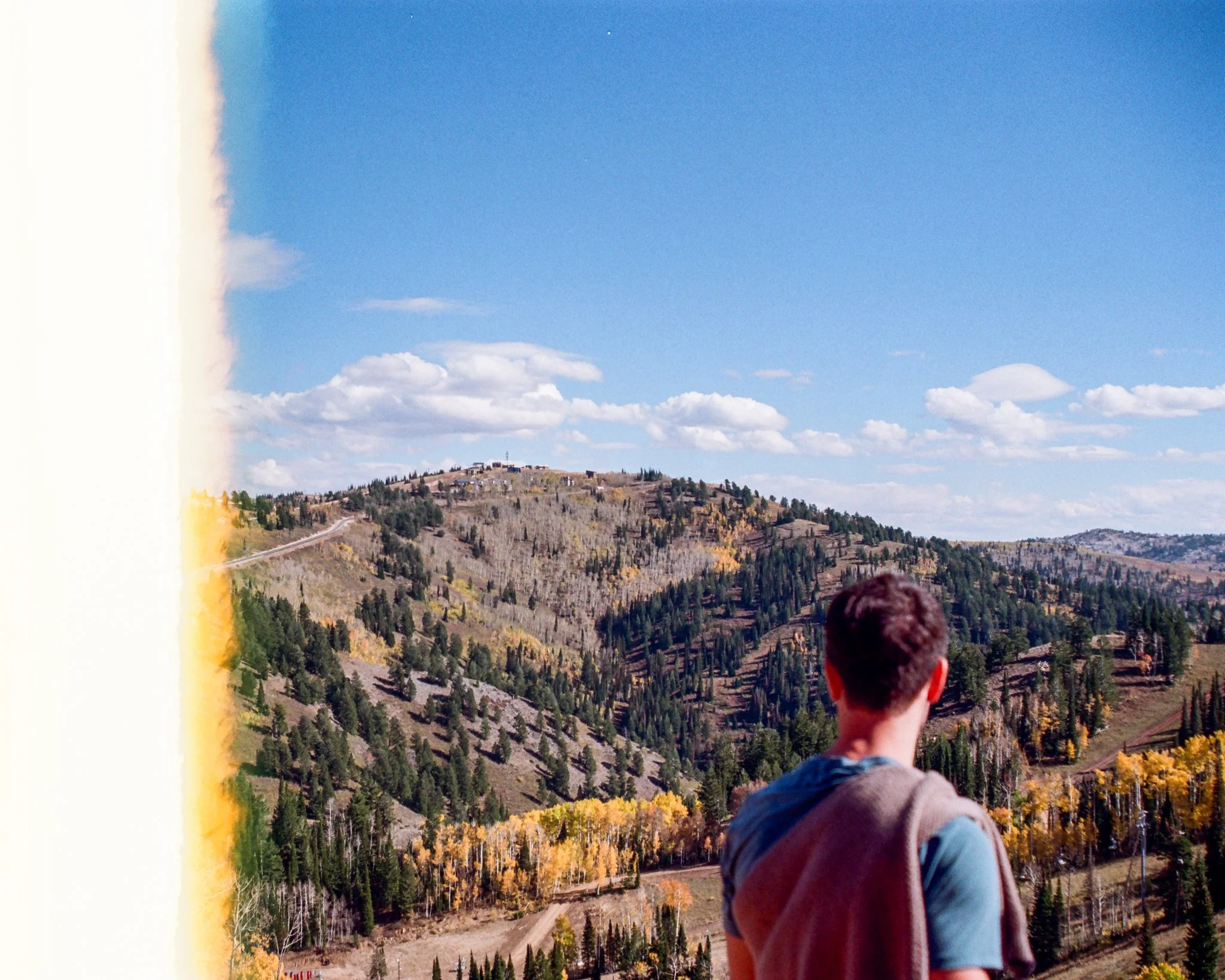 A person standing outdoors looking at a mountainous landscape with trees, hills, and a blue sky with clouds.