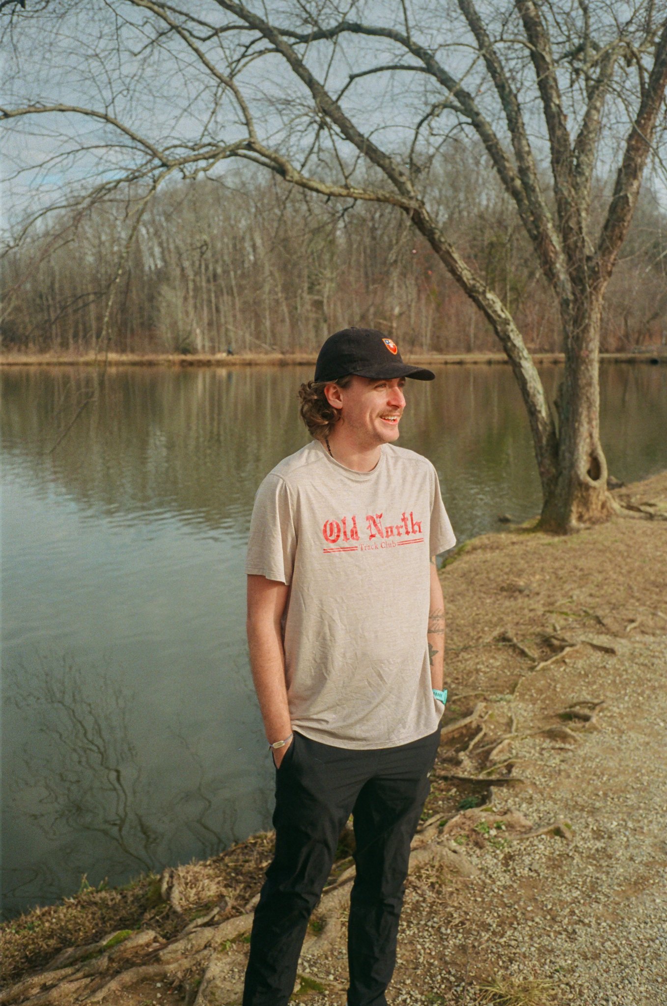 A man with curly hair wearing a black baseball cap, gray t-shirt with red writing, dark pants, and a wristband stands by a lake with trees in the background, smiling and looking to the side.