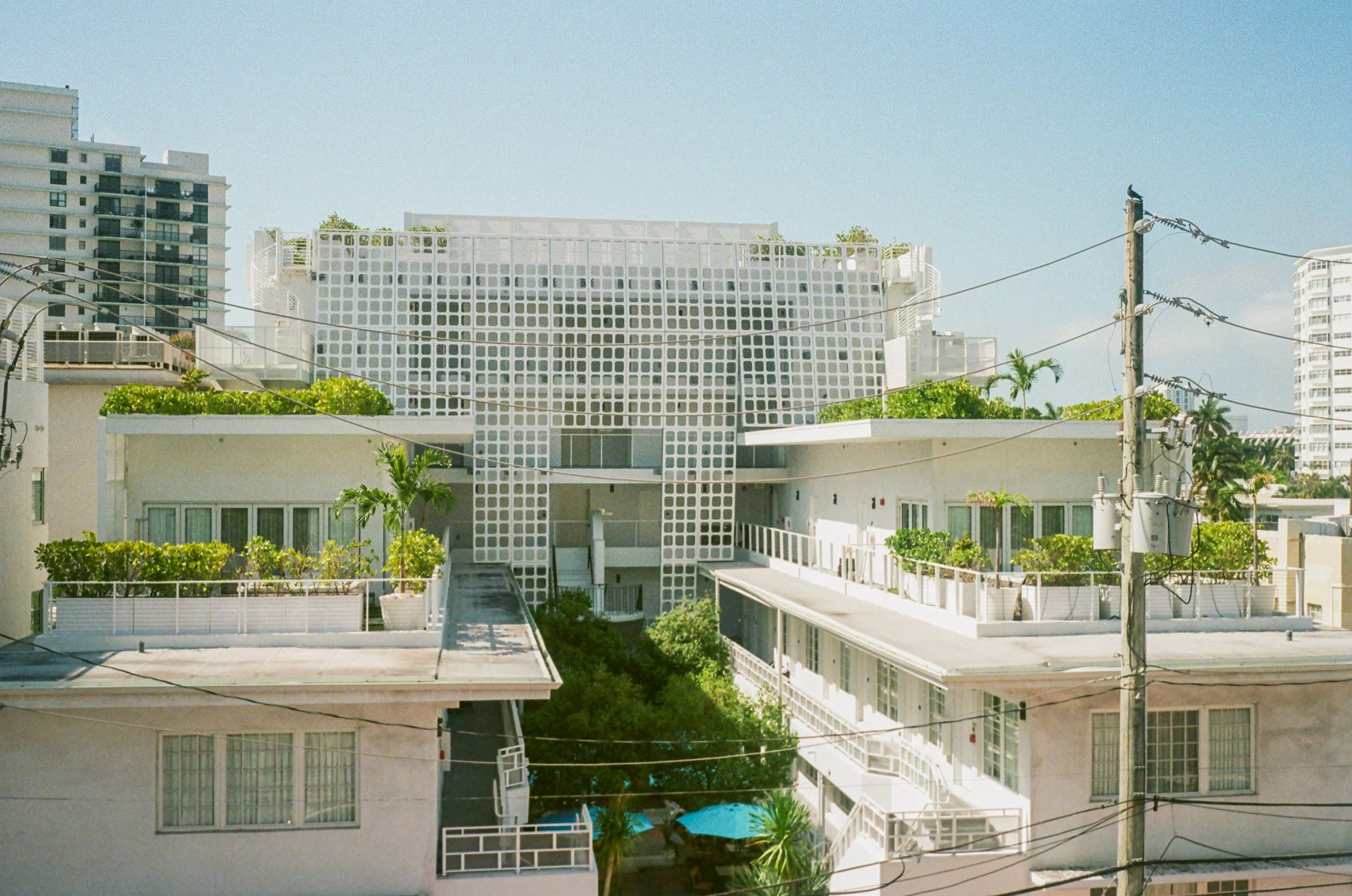 View of modern white buildings with green rooftop gardens, utility poles with wires, and tall apartment buildings in the background under a blue sky.