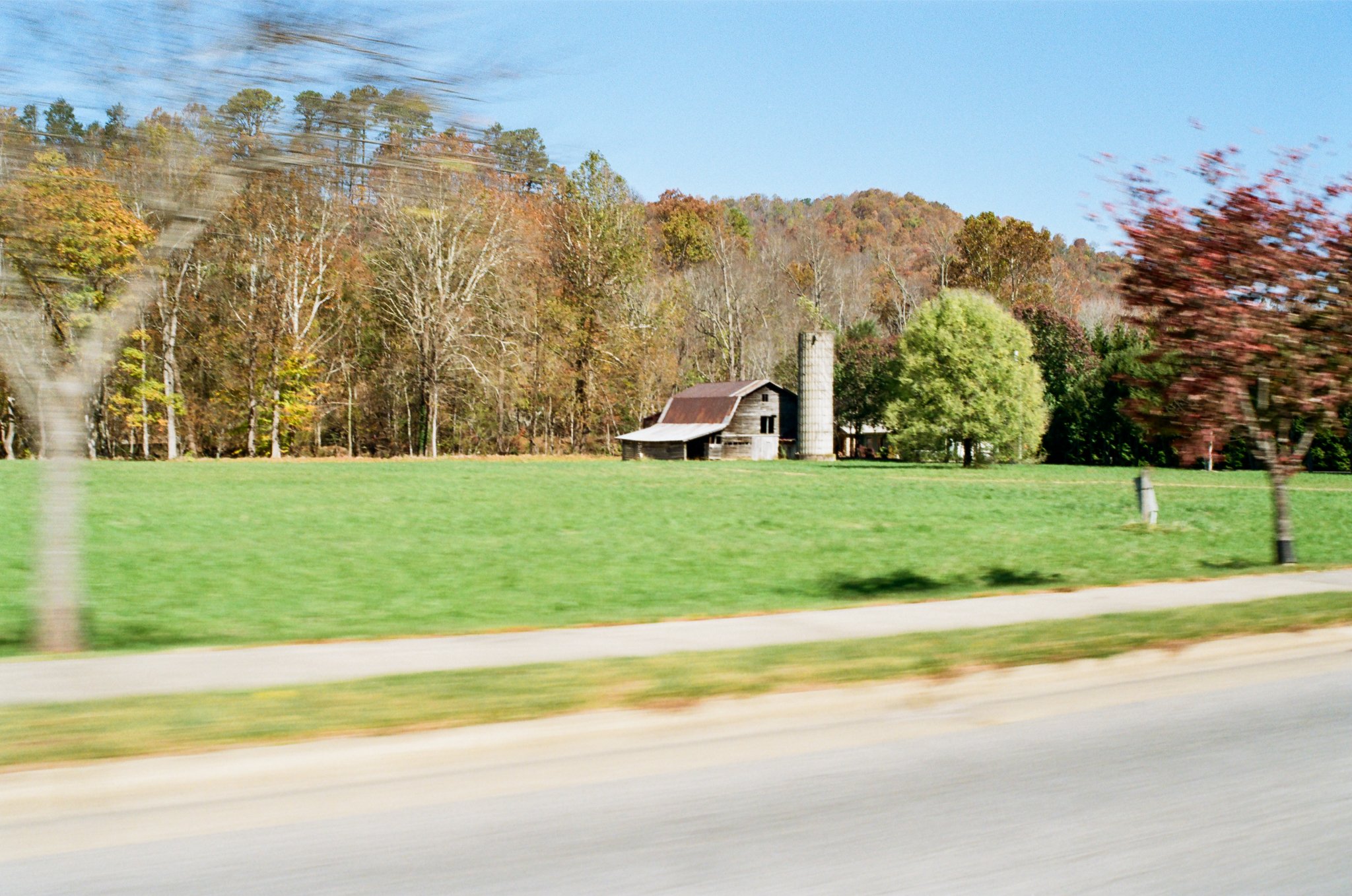 A rural landscape with a barn, silo, and trees in Autumn, seen from a moving vehicle.