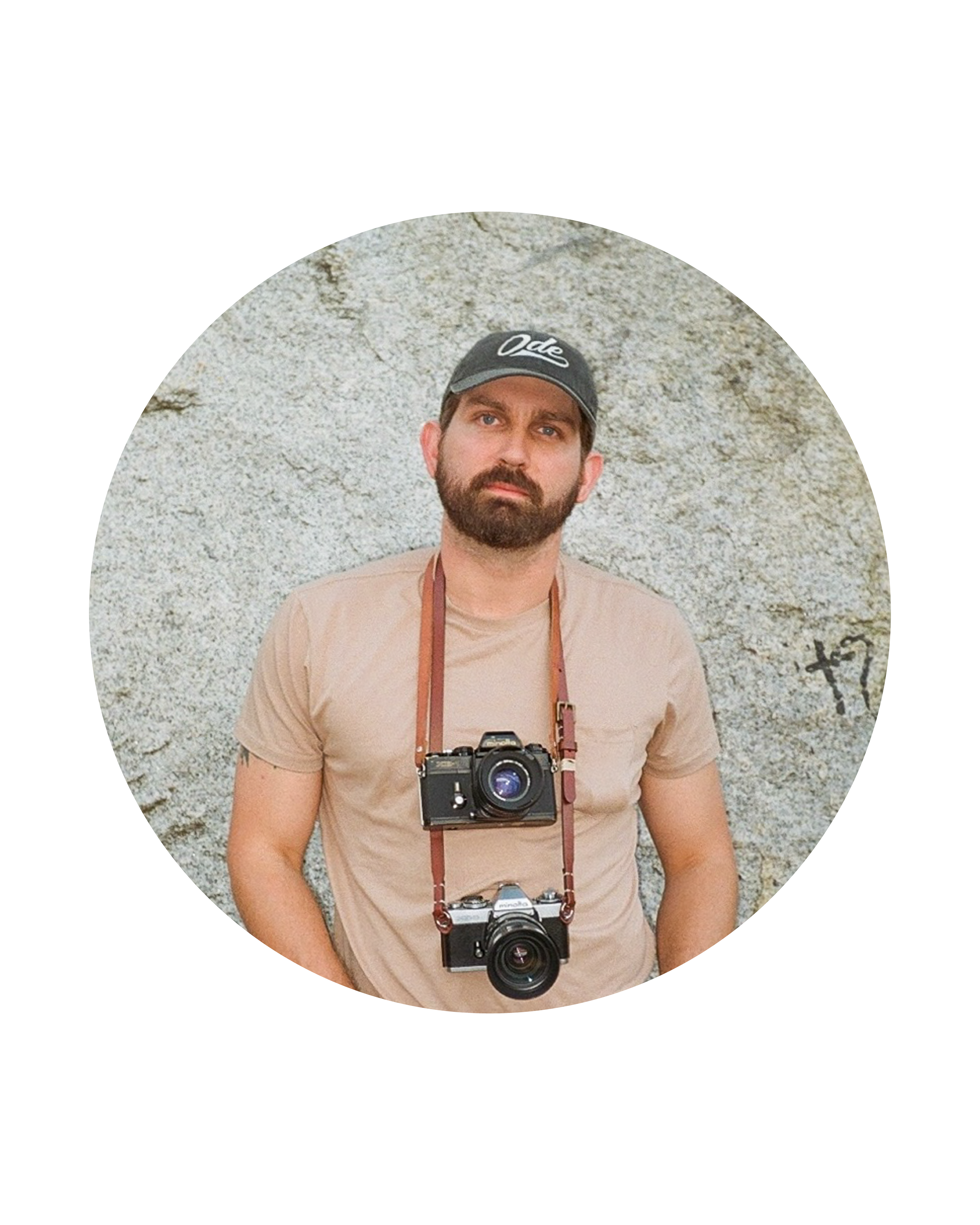 The photographer Connor Poore with a beard wearing a baseball cap and a beige t-shirt with two cameras hanging around his neck, standing in front of a textured stone wall.