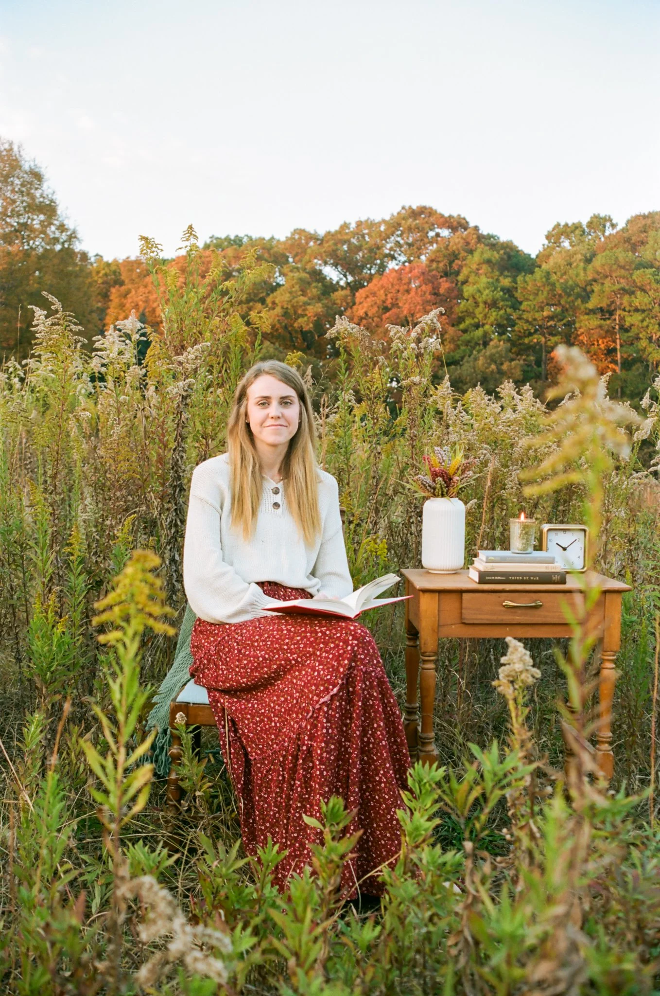 A woman sitting on a chair in a field with a small wooden table beside her, holding an open book, with autumn trees in the background.