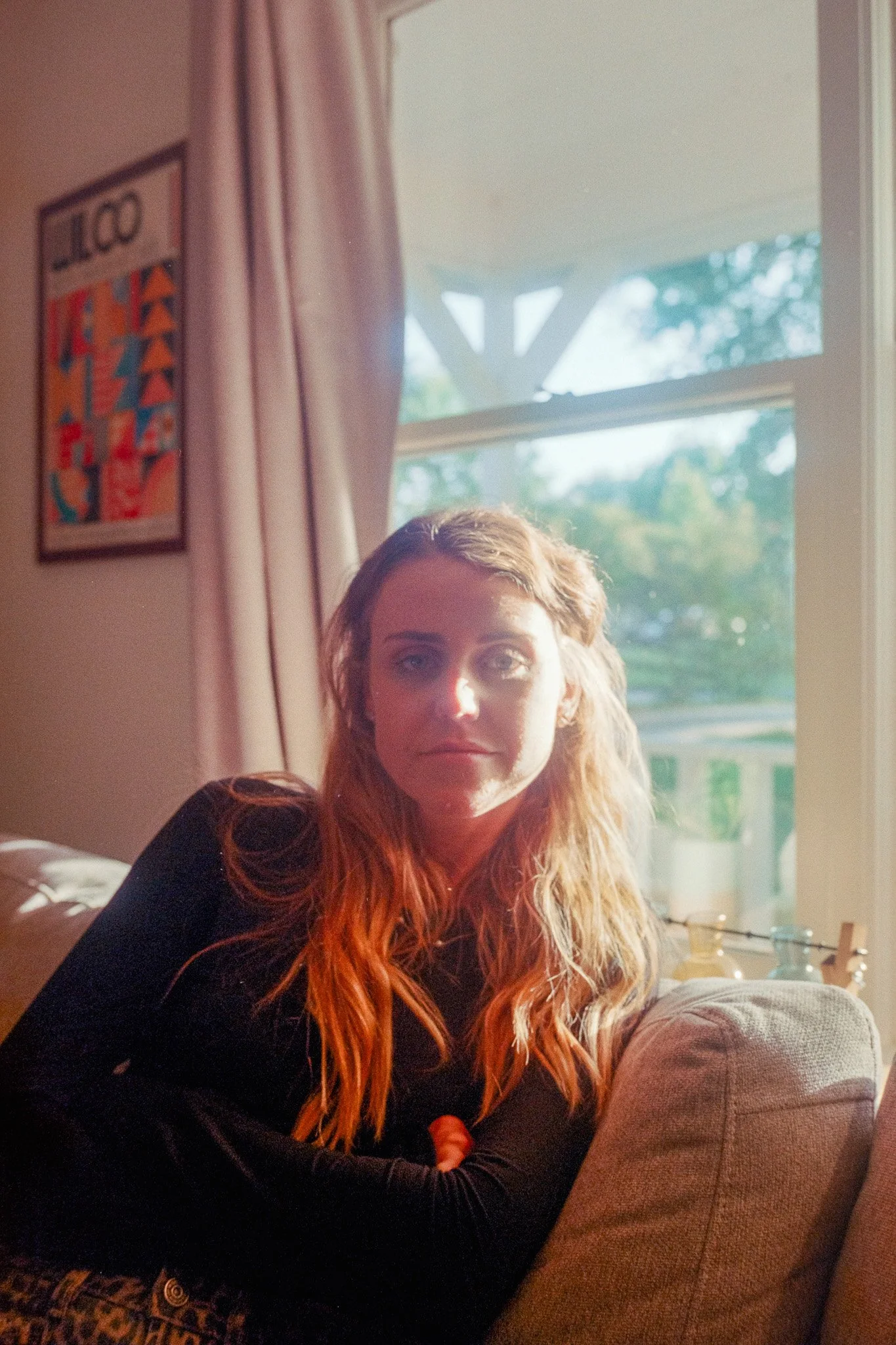 A woman with long wavy red hair sitting on a sofa in a well-lit room, with sunlight coming through a large window behind her.