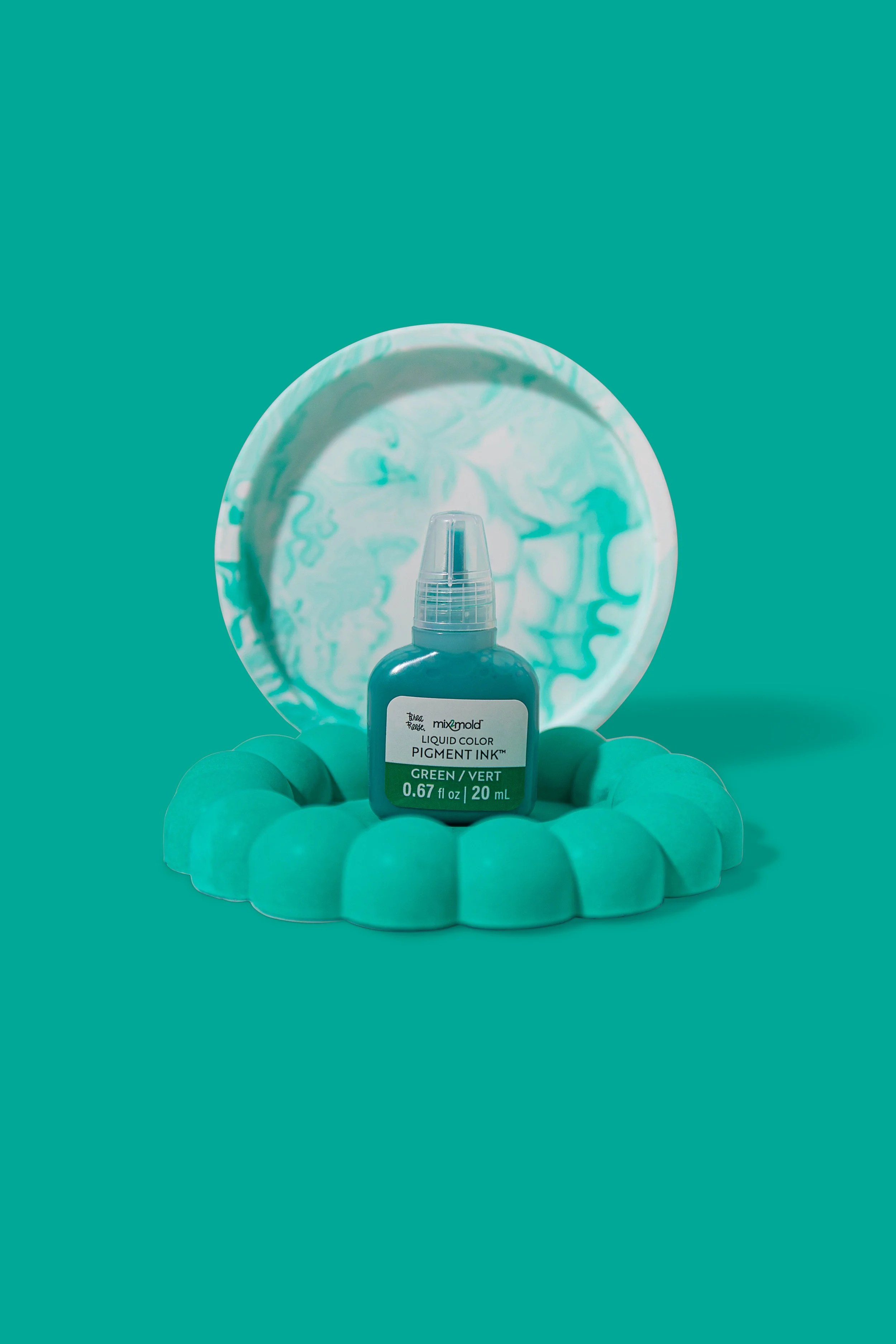 A teal-colored bottle of liquid color pigment ink labeled 'Green' from the brand 'mix mold' placed on a teal oval base with a mirror behind it reflecting a swirling pattern in the same color.
