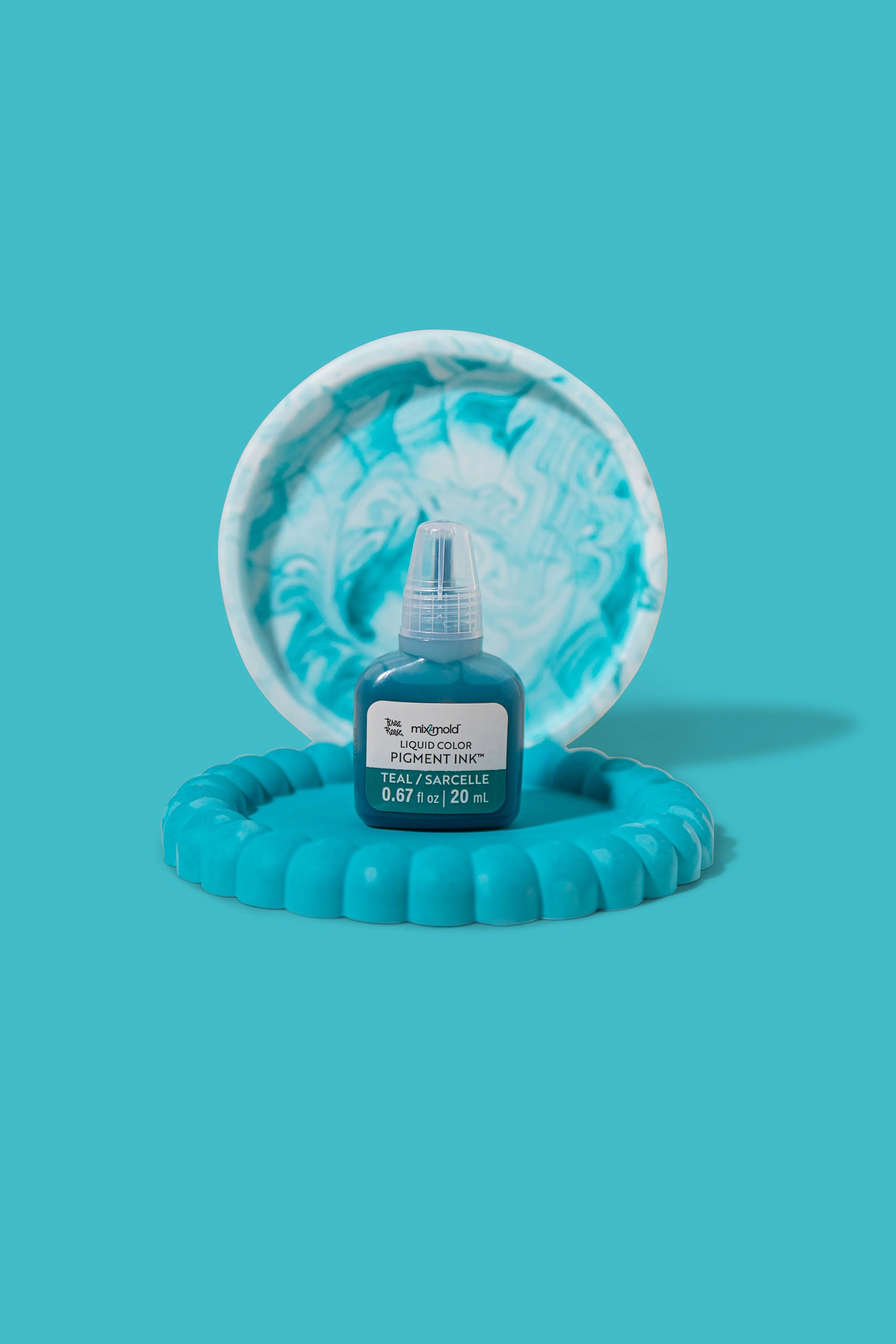 A teal liquid pigment ink bottle on a blue textured circular tray with a marble-patterned mirror behind it.