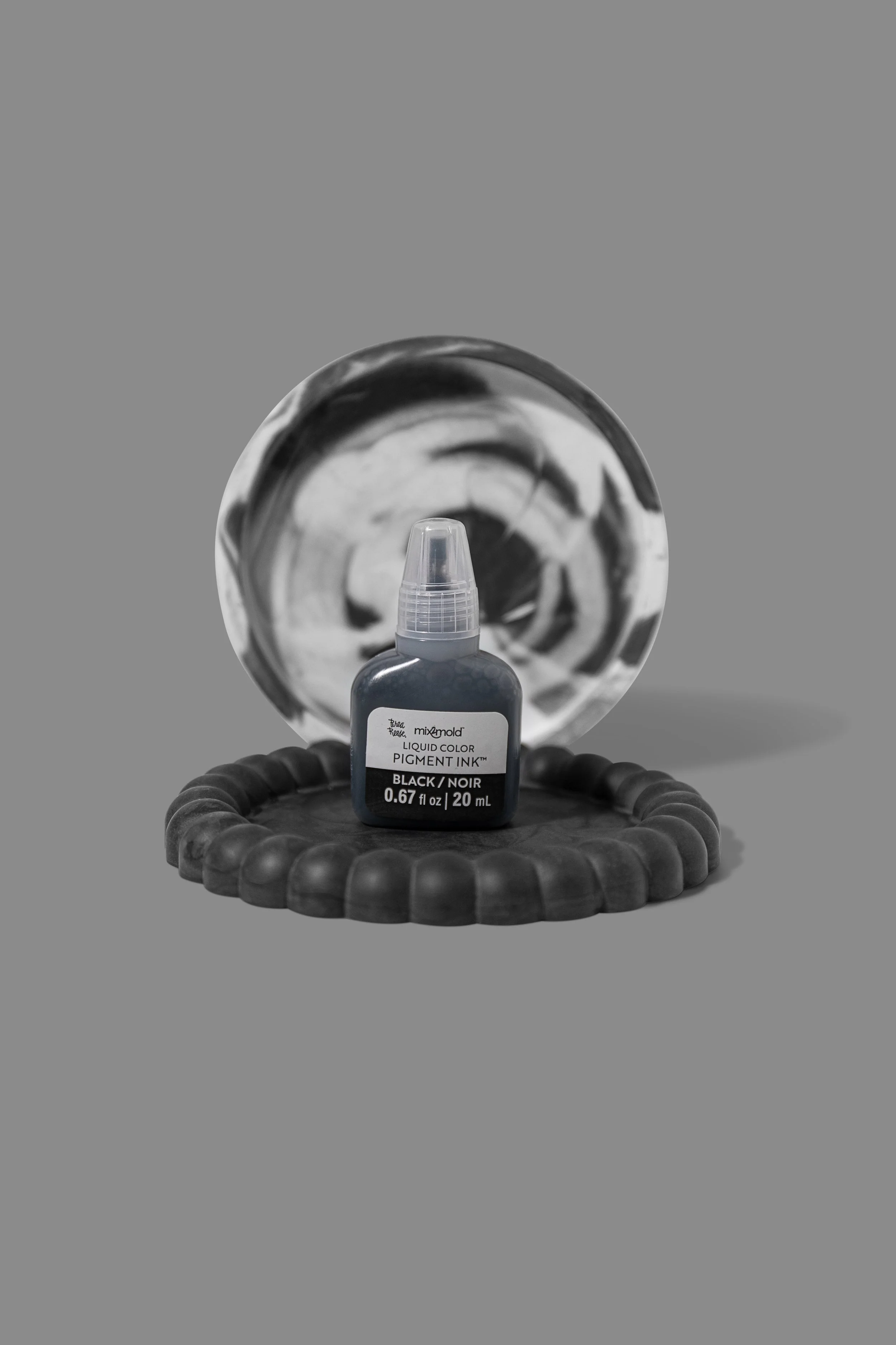 A bottle of black liquid pigment ink, placed on a black coiled object, with a round patterned background and a reflective surface.