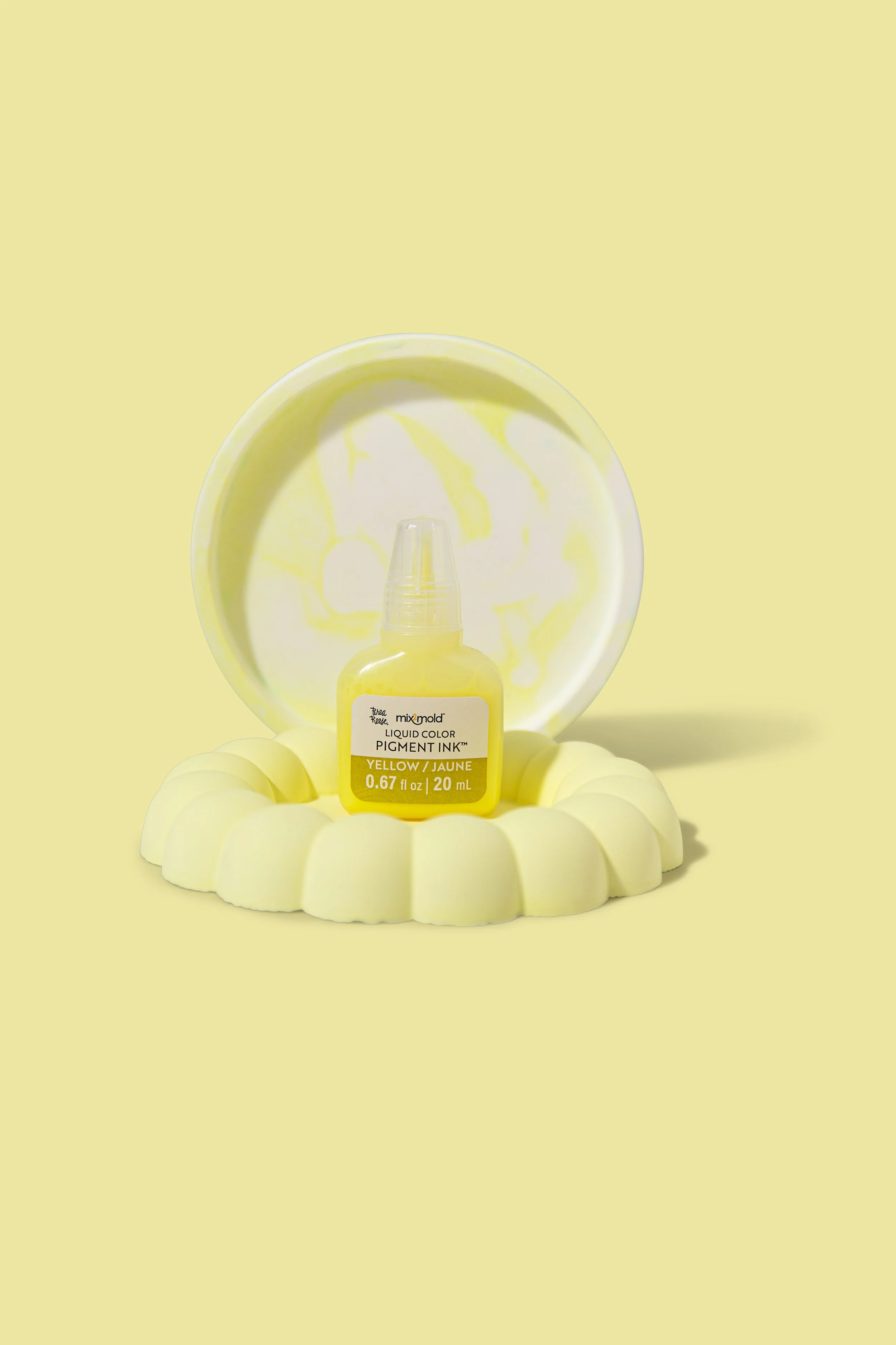 A bottle of yellow liquid pigment ink placed on a yellow circular platform with a scalloped edge, set against a yellow background, with a round yellow container behind it.