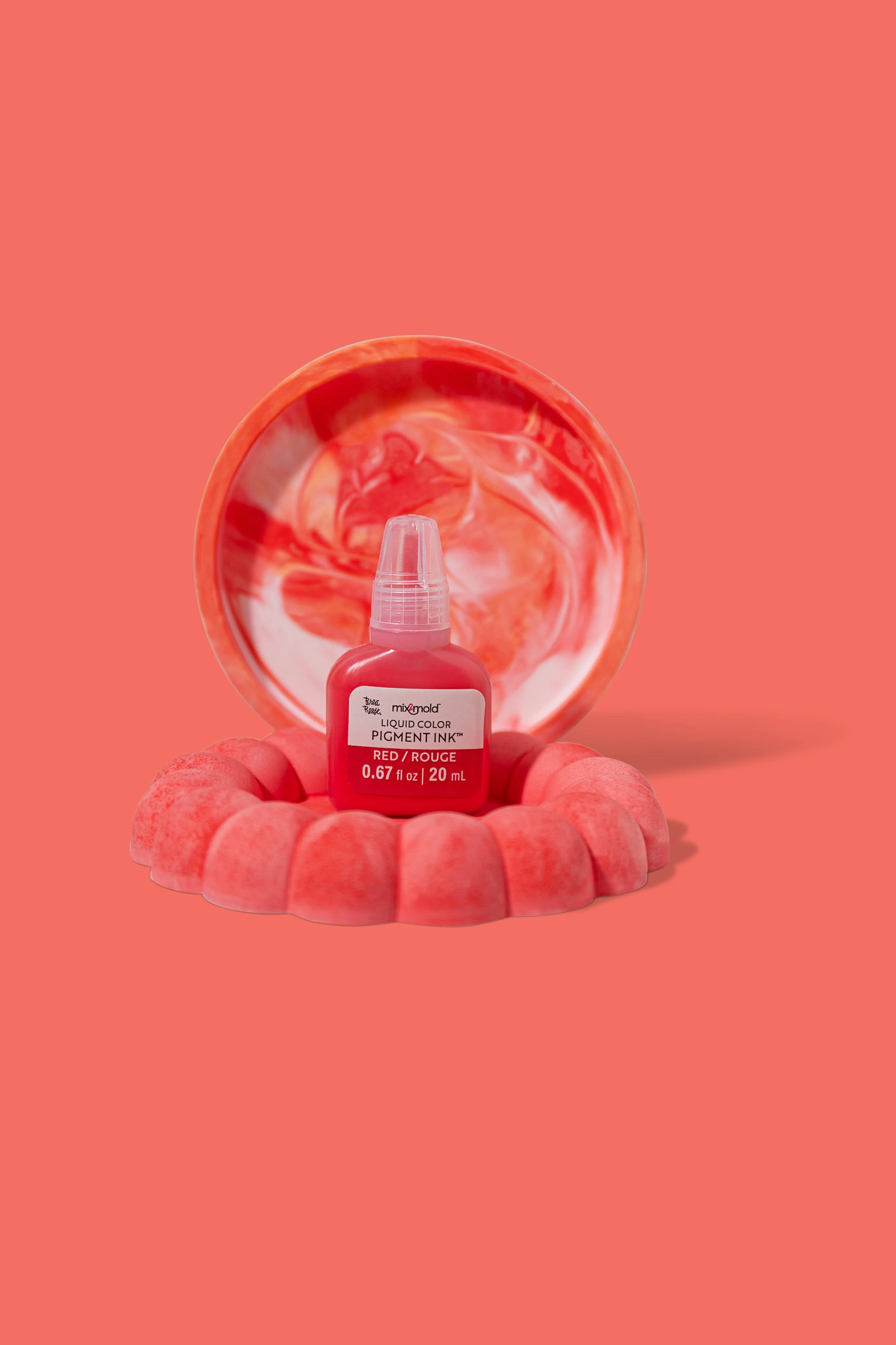 A bottle of liquid red pigment ink is placed on a pink, scalloped stand with a marbled pink and white lid in the background, all against a coral pink backdrop.