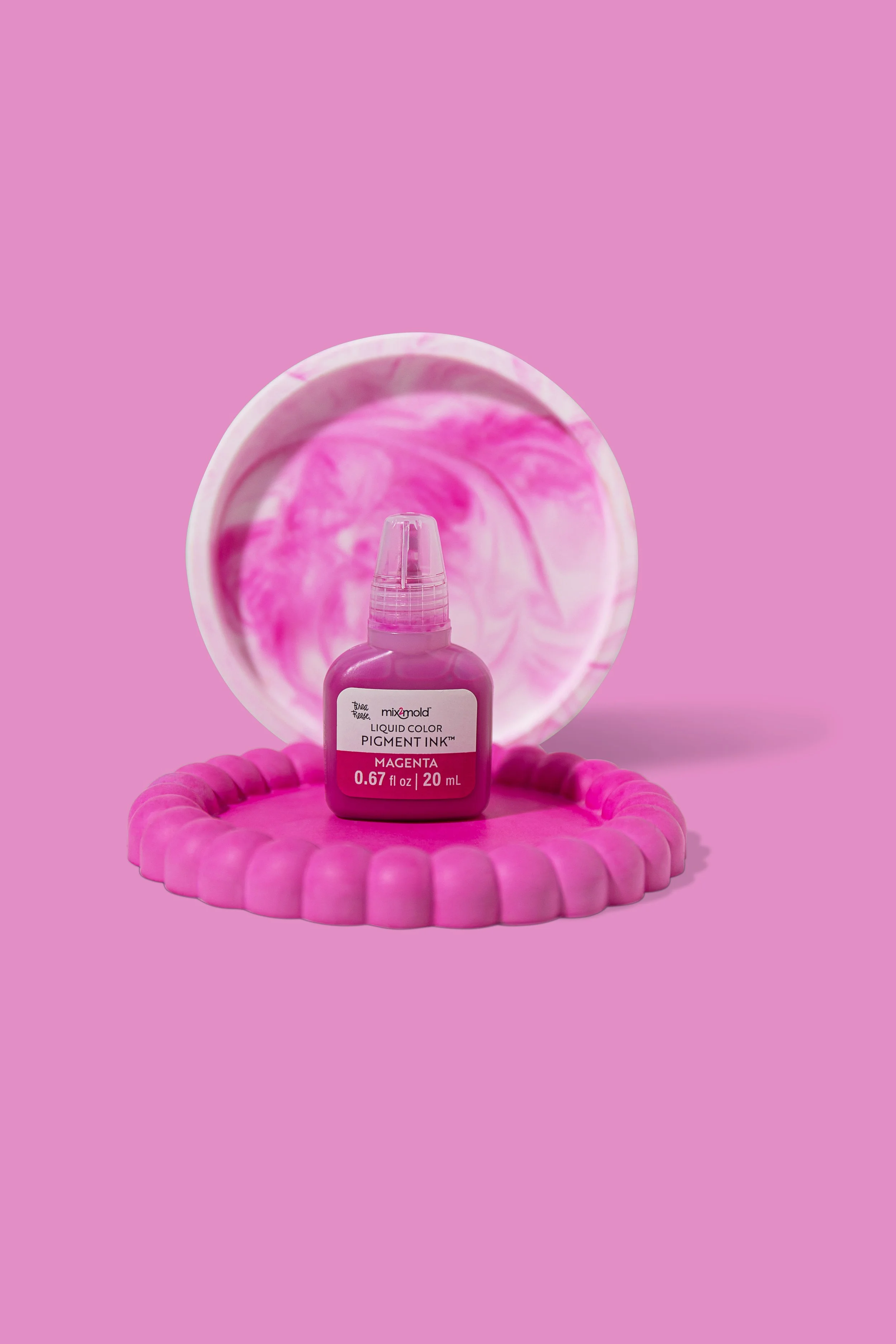A bottle of magenta liquid color pigment ink placed on a pink silicone mat in front of a marble-patterned dish, with a pastel pink background.