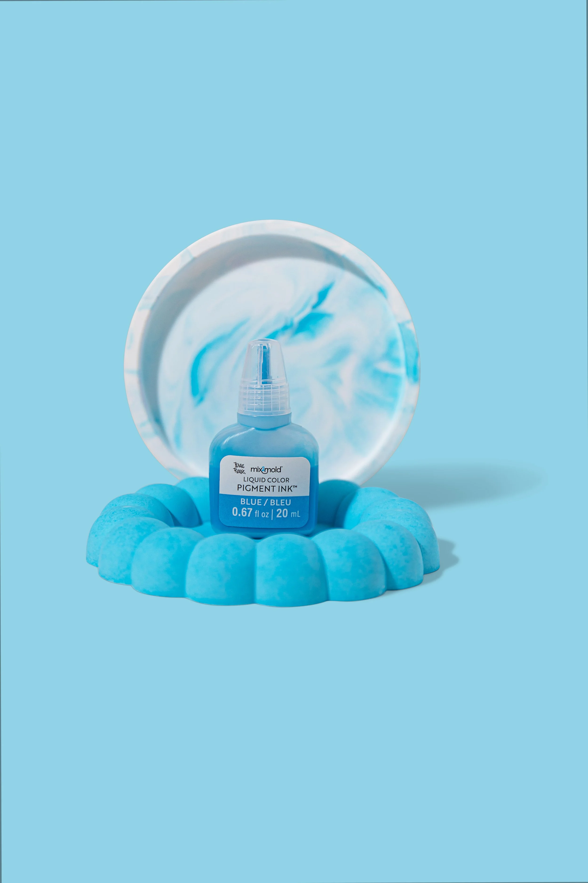 A bottle of blue pigment ink placed on a circular blue foam ring, with a marble-patterned dish behind it, all set against a blue background.