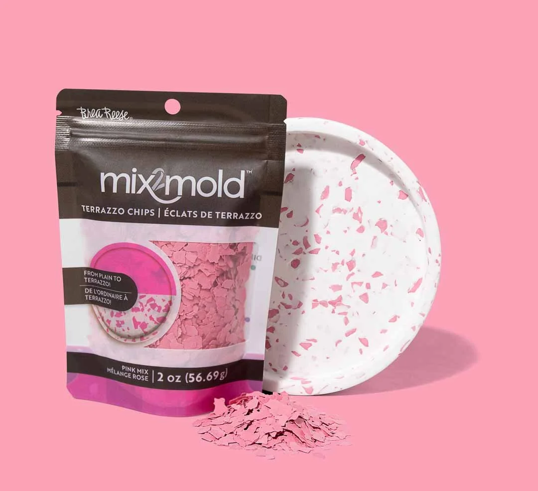 A black and pink package of Mix/Mold terrazzo chips labeled 'Pink Mix' and 'Mélange Rose'. Next to the package, there is a small white terrazzo chip disc with pink chips, and a few pink terrazzo chips spilled on the pink background.
