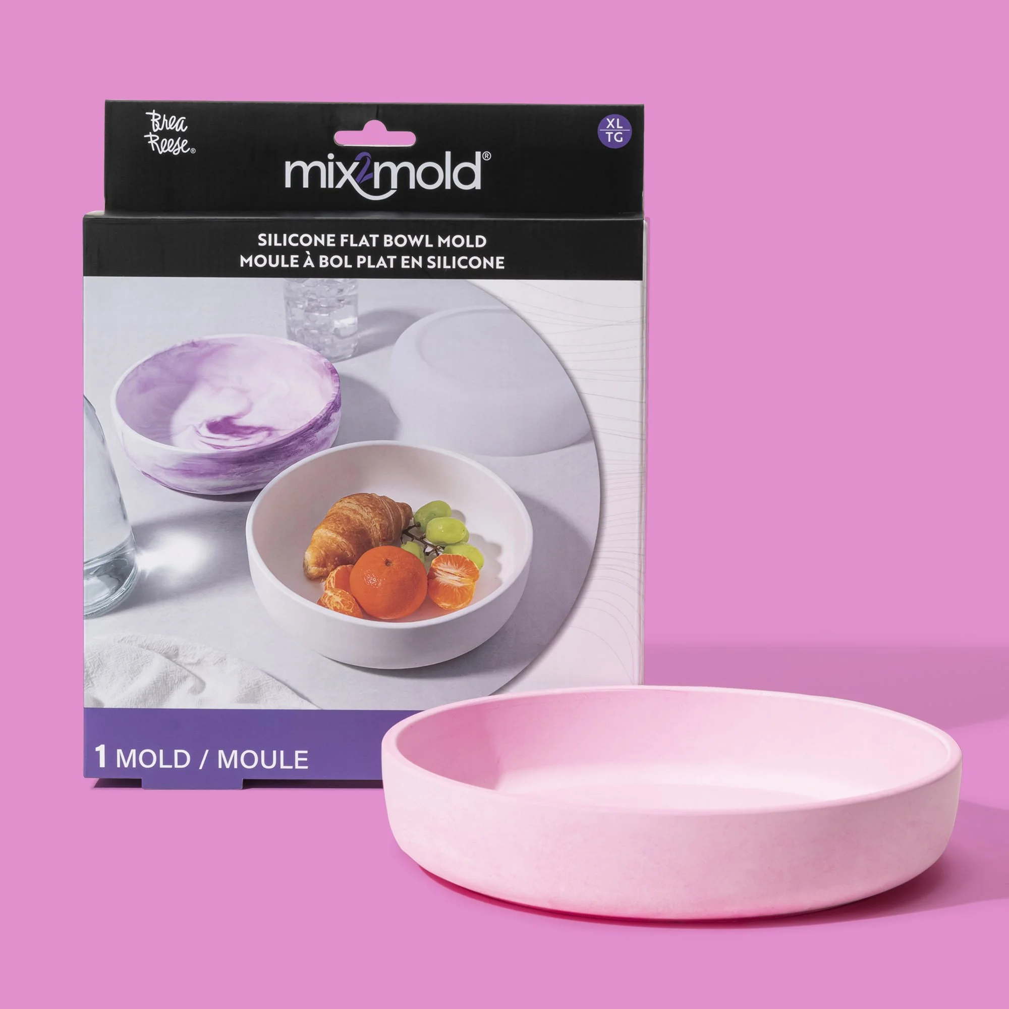 A pink silicone flat bowl mold placed in front of its packaging box, which displays an image of the same bowl holding various fruits and a purple marbled bowl.