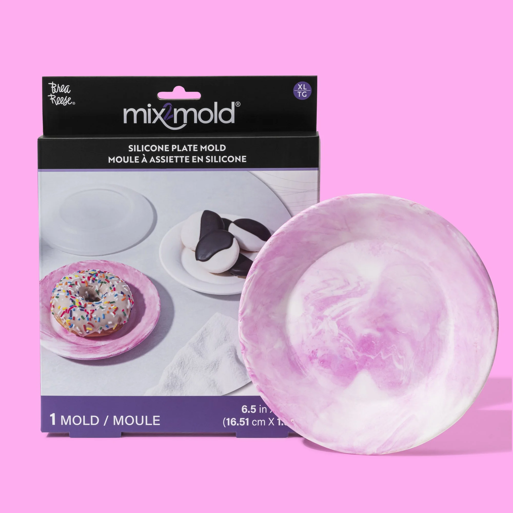 A pink and white marbled silicone plate mold next to the packaging, which is black with purple and white details, showing an example of a white plate with a donut and some chocolates.