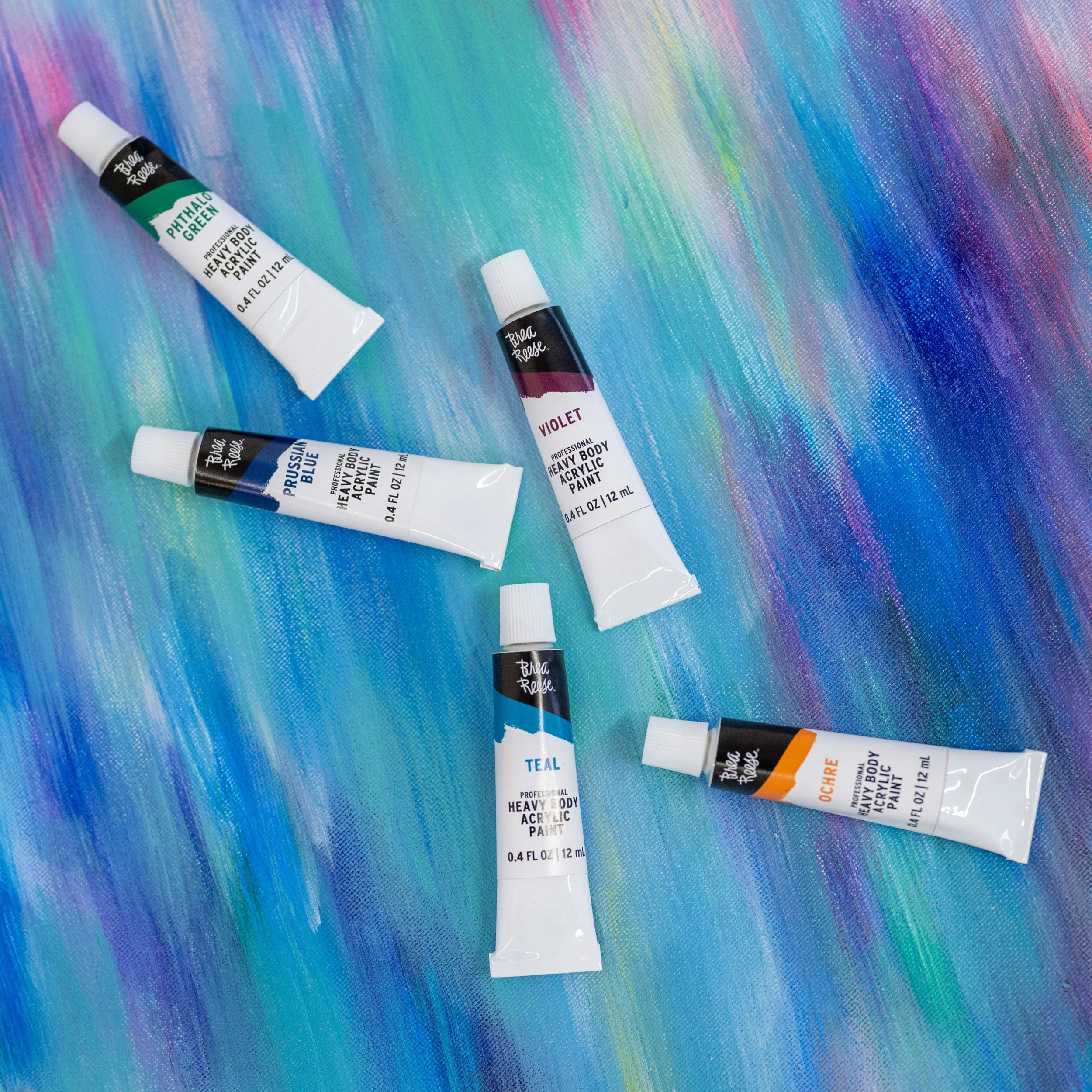 Five tubes of heavy body acrylic paint in different colors: phthalo green, violet, prussian blue, teal, and ochre, arranged on a colorful, textured canvas background.